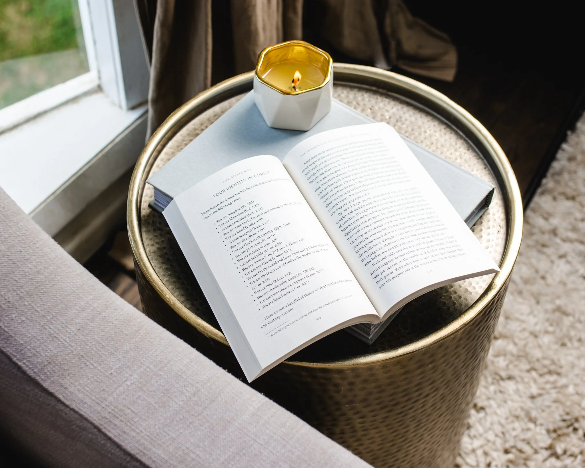 Open book resting on a round, gold-trimmed table, with a white candle in a hexagonal holder beside it, near a window letting in natural light.