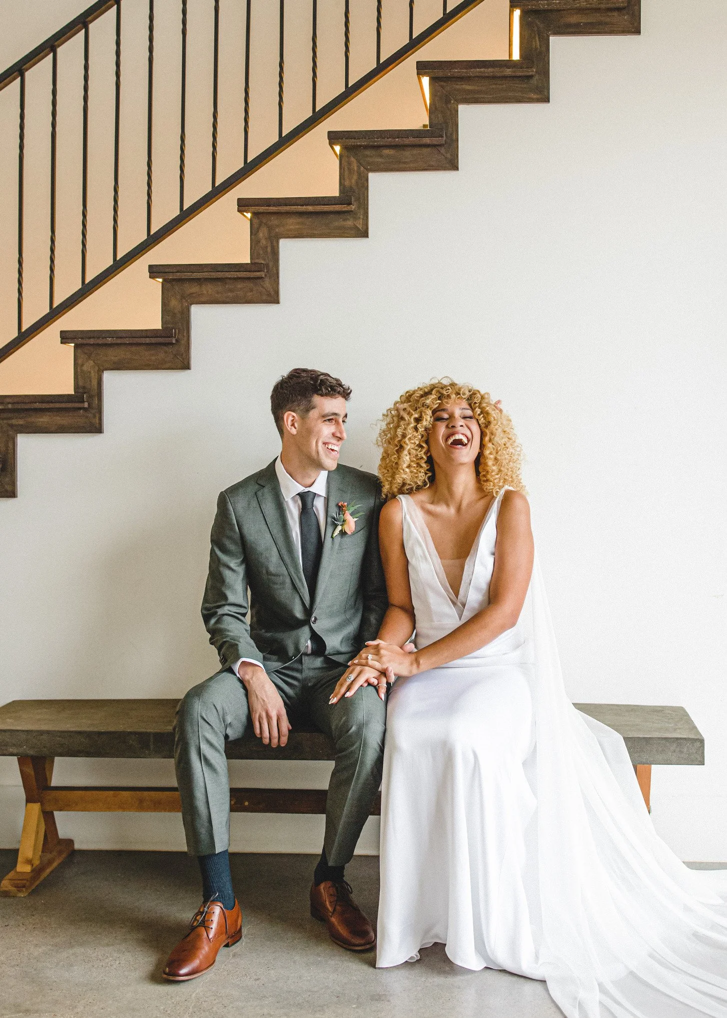 A couple in wedding attire sitting on a wooden bench underneath a staircase, smiling and laughing.
