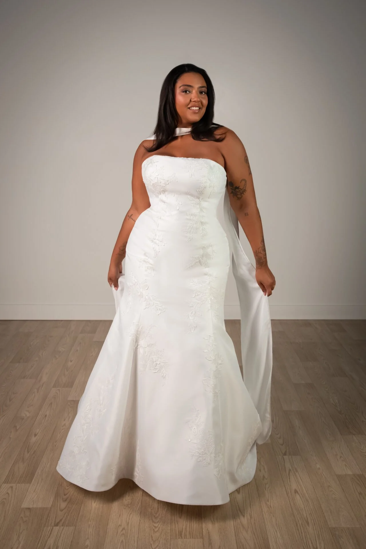 A woman wearing an ivory Stella York wedding dress with floral embroidery standing on a wooden floor against a plain light gray wall.