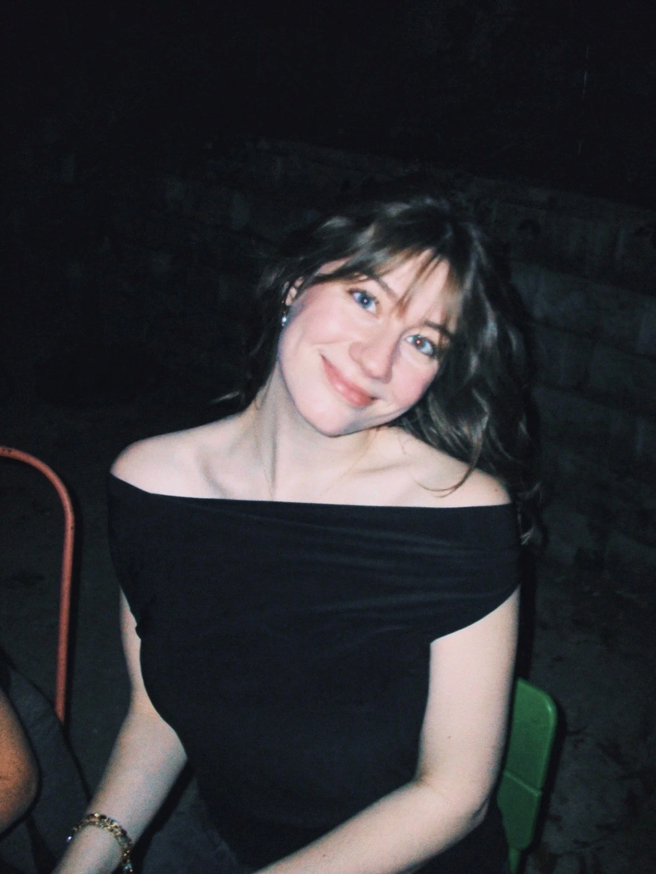 A woman with black, wavy hair and blue eyes, smiling, sitting in a dark environment, wearing a black off-the-shoulder dress.
