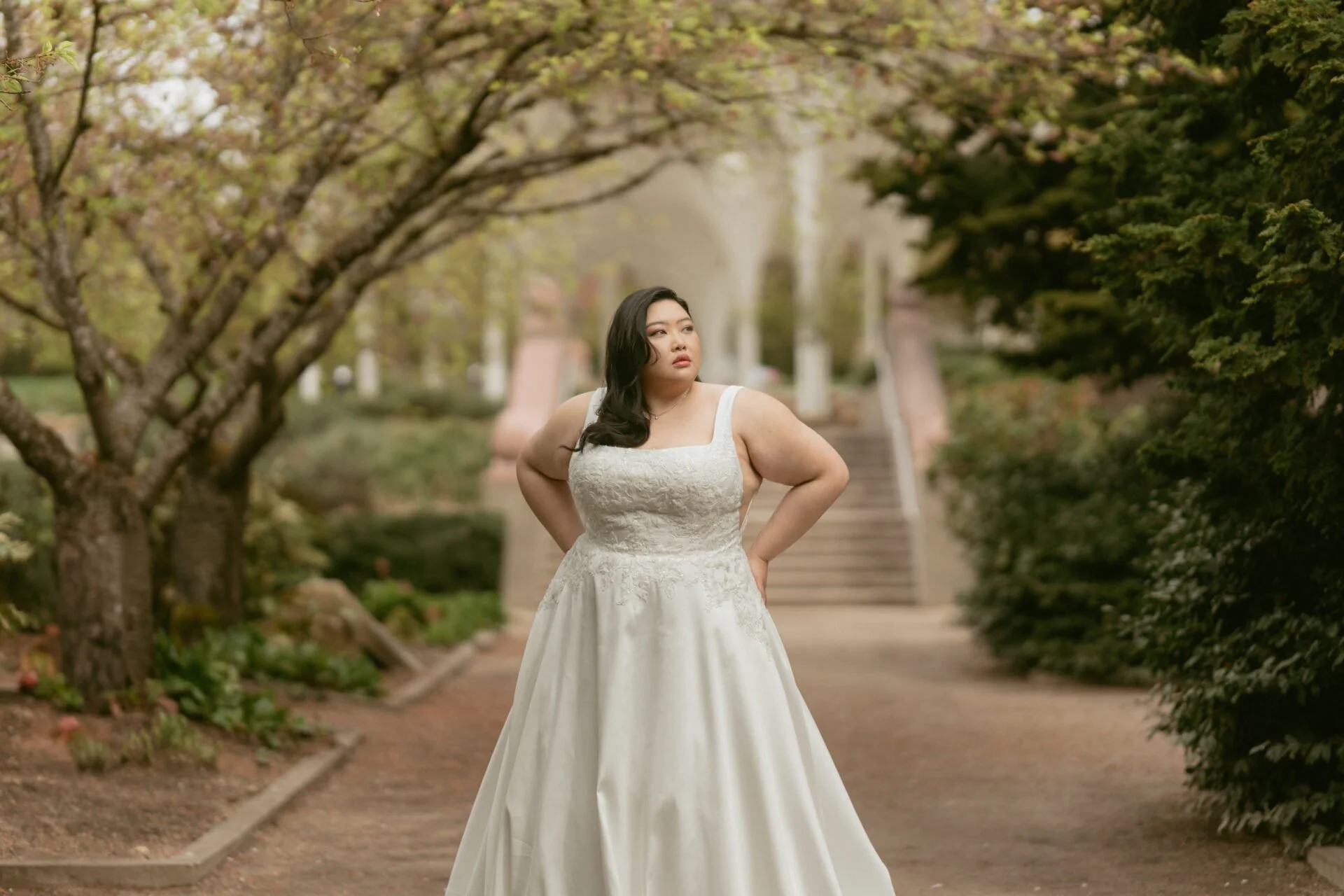A woman wearing an ivory, satin, lace embellished, square neck Stella York wedding dress standing in a park surrounded by trees with light green and brown leaves.