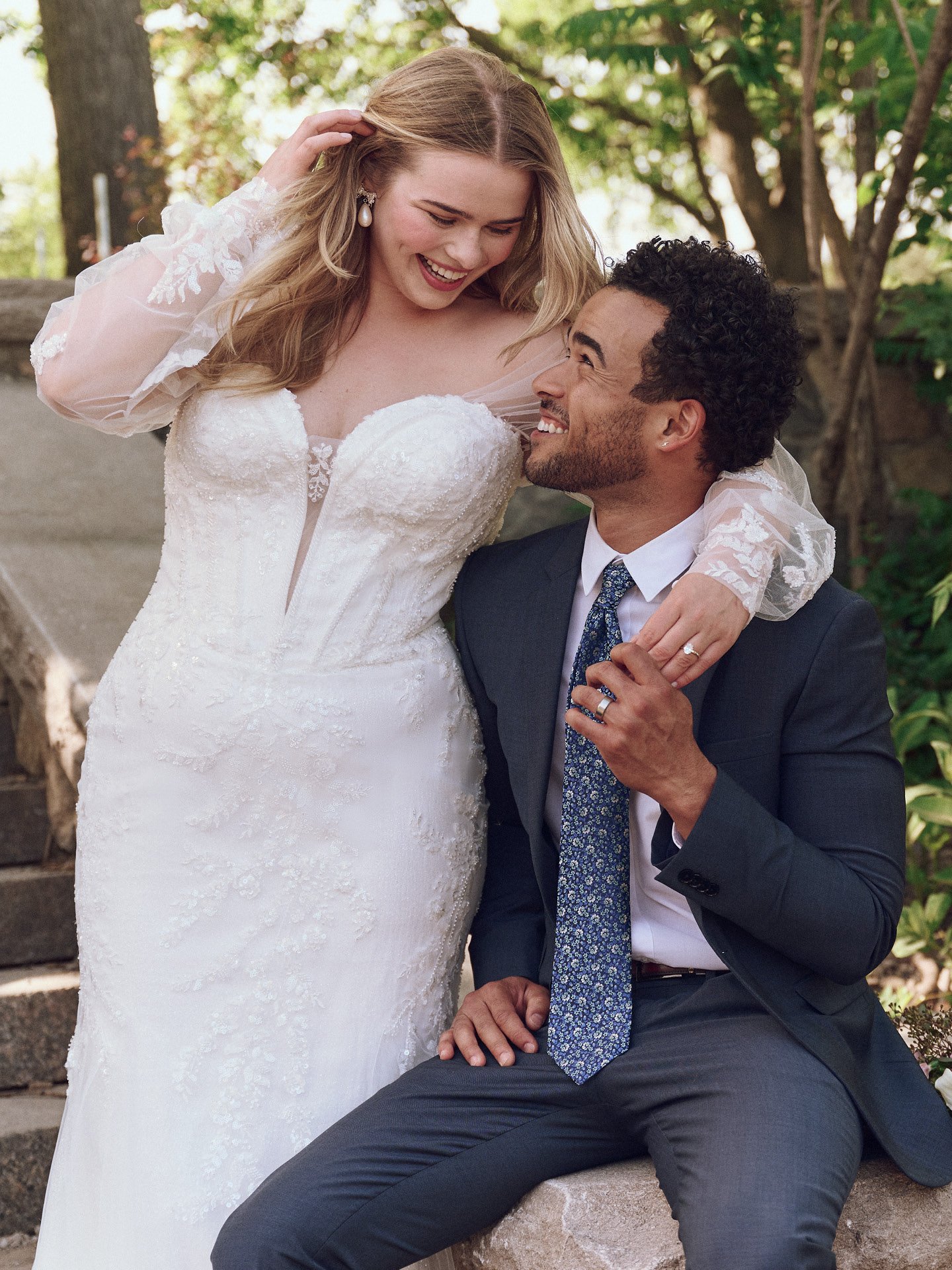 A bride and groom smiling and looking at each other outdoors. The bride is in a Maggie Sottero gown with lace, sparkle, and a plunging sweetheart neckline.