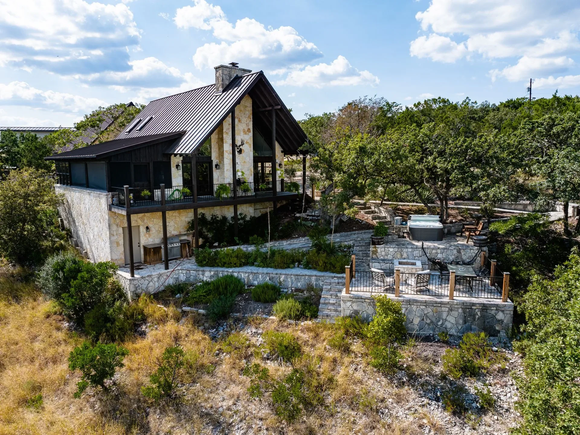 A modern house with a high, steep metal roof, stone walls, large windows, and a spacious outdoor patio area surrounded by trees and greenery.