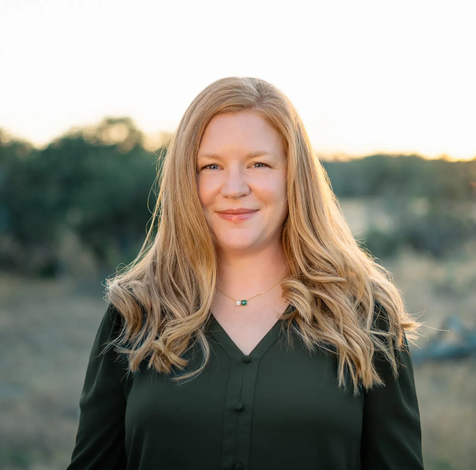 Portrait of a woman with long, curly blonde hair smiling outdoors during sunset, wearing a dark green top and a necklace with a green and white pendant.
