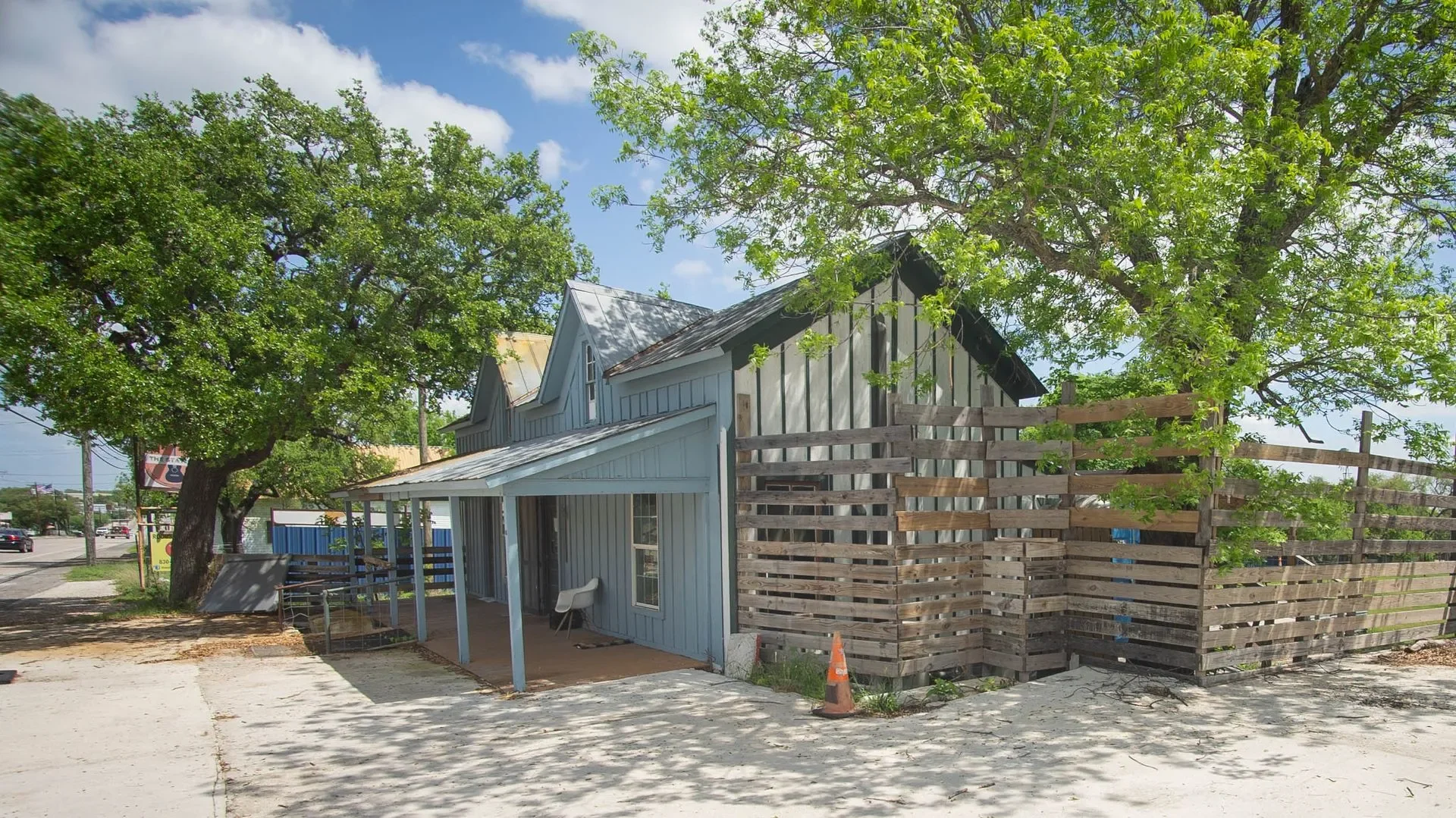 A blue house with a metal roof under large leafy trees on a sunny day, with a wooden fence and sidewalk in front.