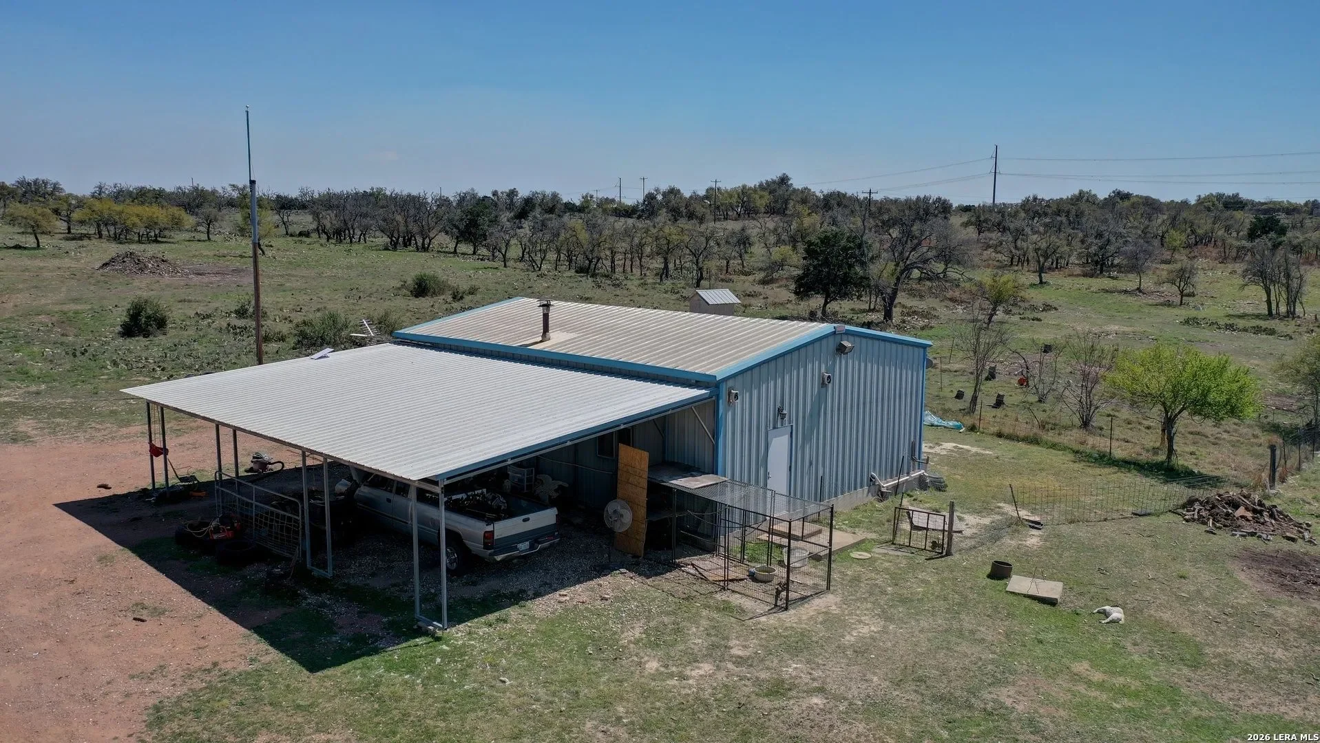 A rural scene with a metal building featuring a covered carport, parked vehicle, and various tools and equipment, set against a backdrop of sparse trees and open land under a clear blue sky.