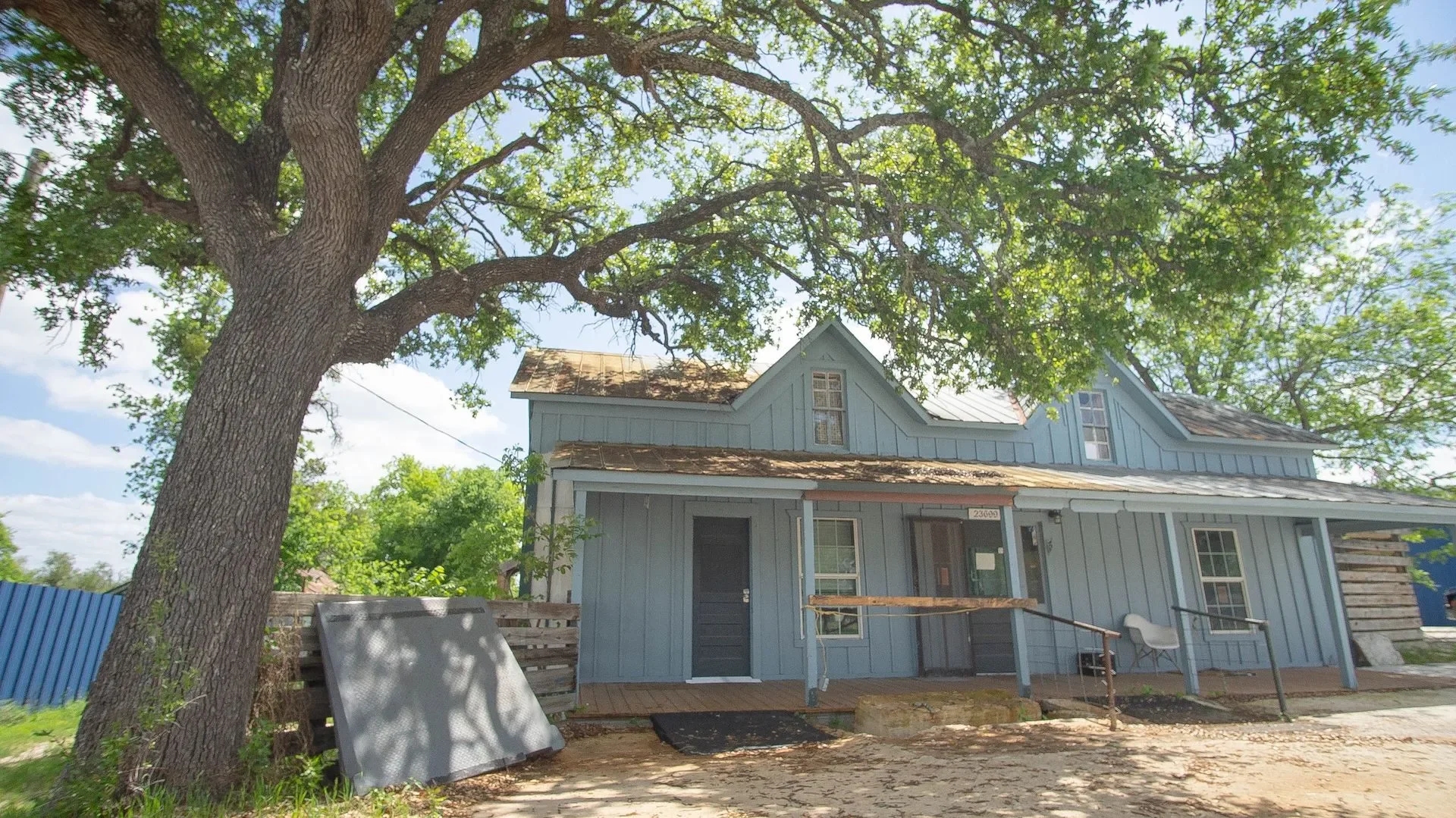 A light blue house with a gabled roof and a small porch, situated in a yard with a large tree in front. The tree has green leaves and provides shade, while the house appears to be undergoing some renovations.