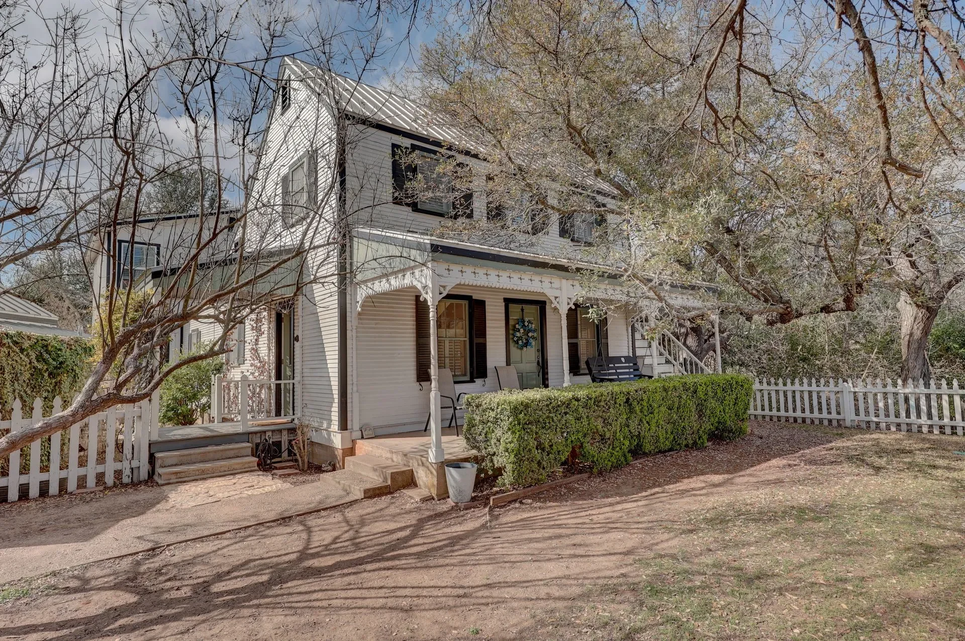 A white two-story house with a porch, black shutters, and a decorative wreath on the door, surrounded by trees and a white picket fence, with a dirt pathway in front.