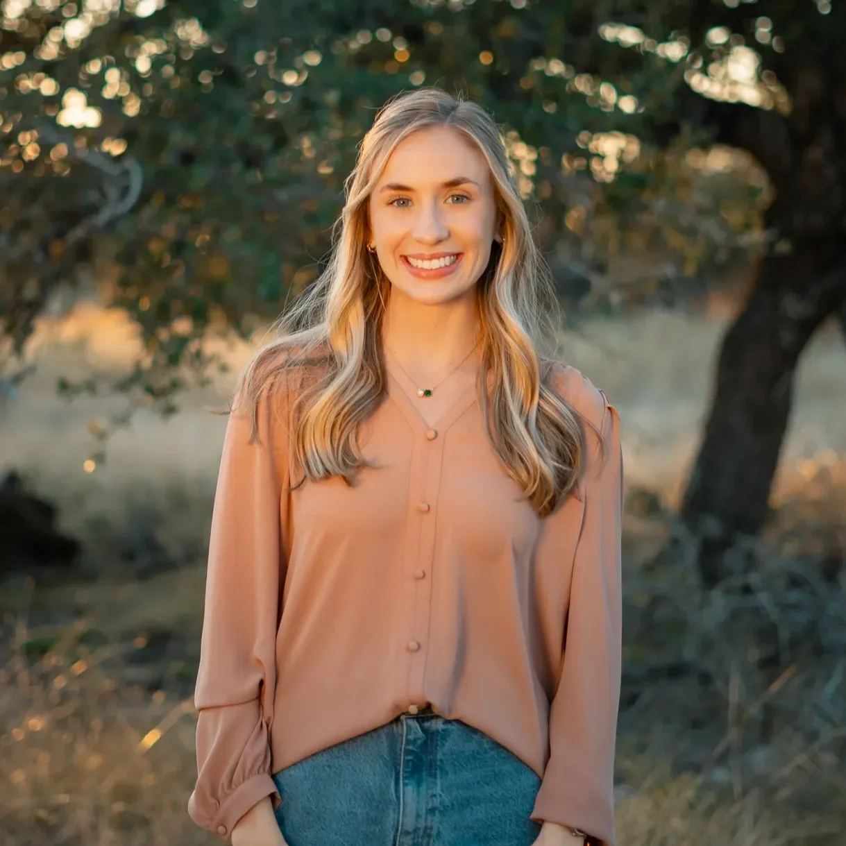 A young woman with blonde hair, wearing a beige blouse and jeans, smiling outdoors during sunset, with trees in the background.