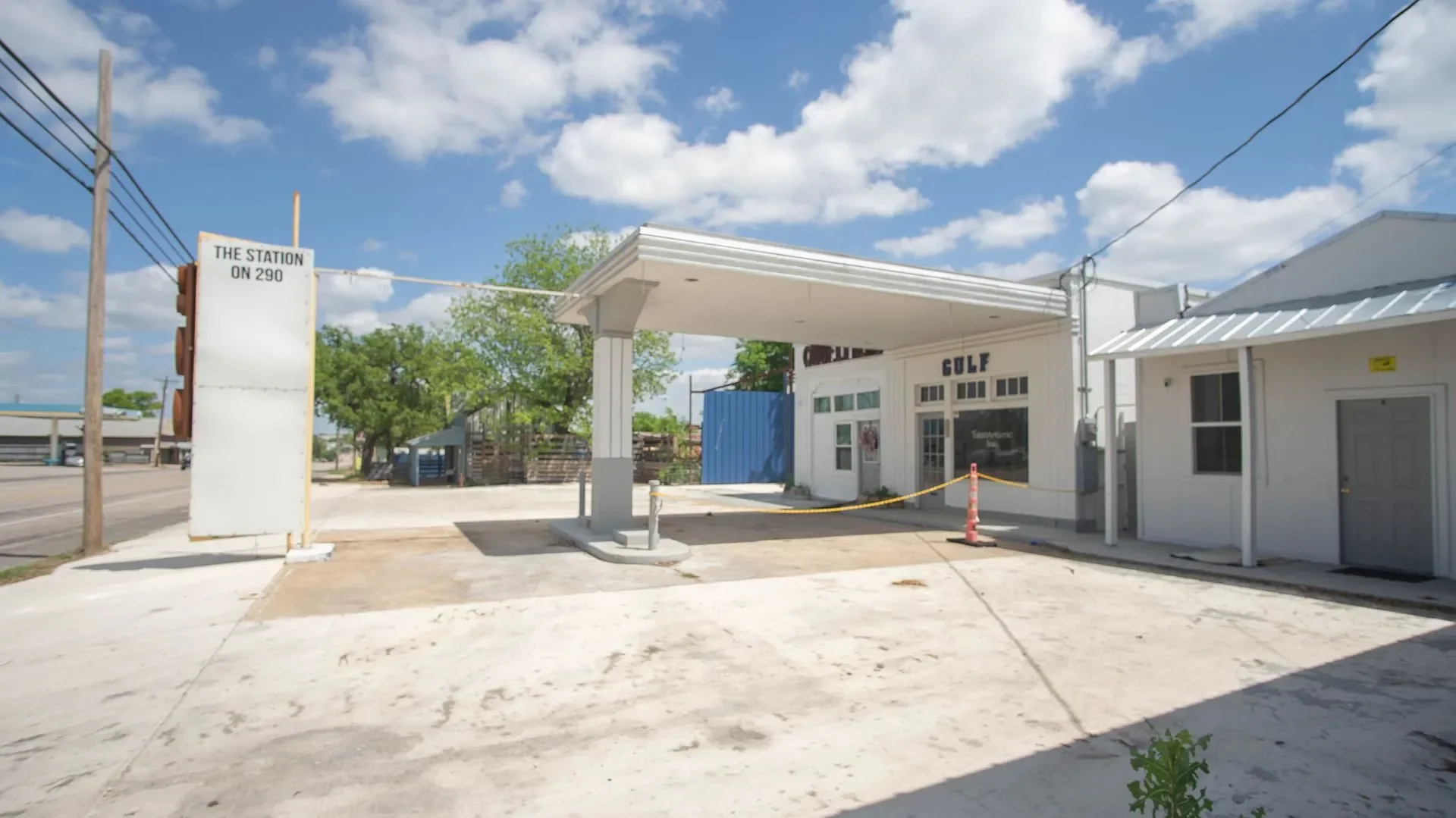 Small roadside gas station with a white building and an awning, a large empty sign, and a road with utility poles, trees, and blue sky with clouds in the background.