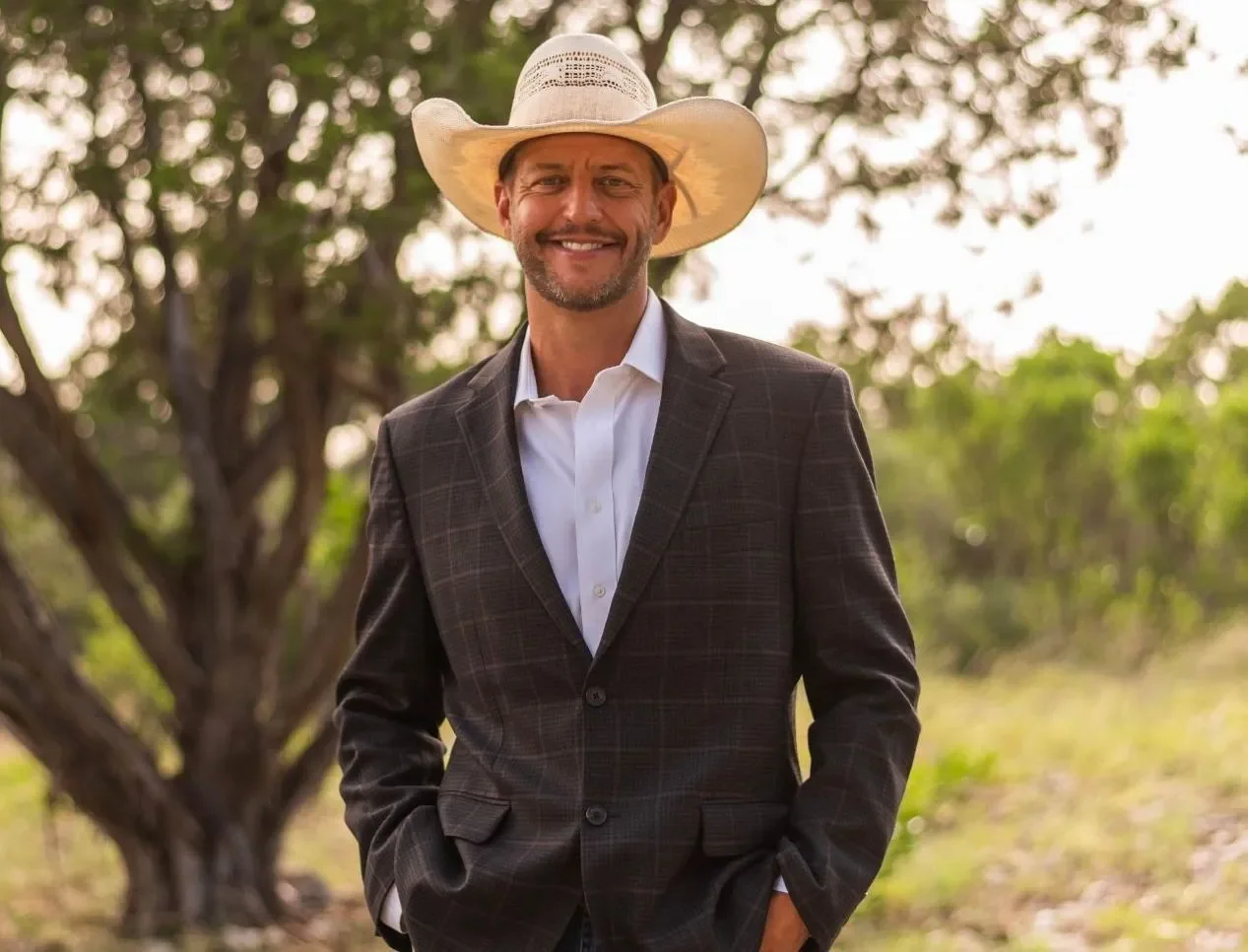 A man wearing a suit and a cowboy hat standing outdoors next to a tree, smiling at the camera.