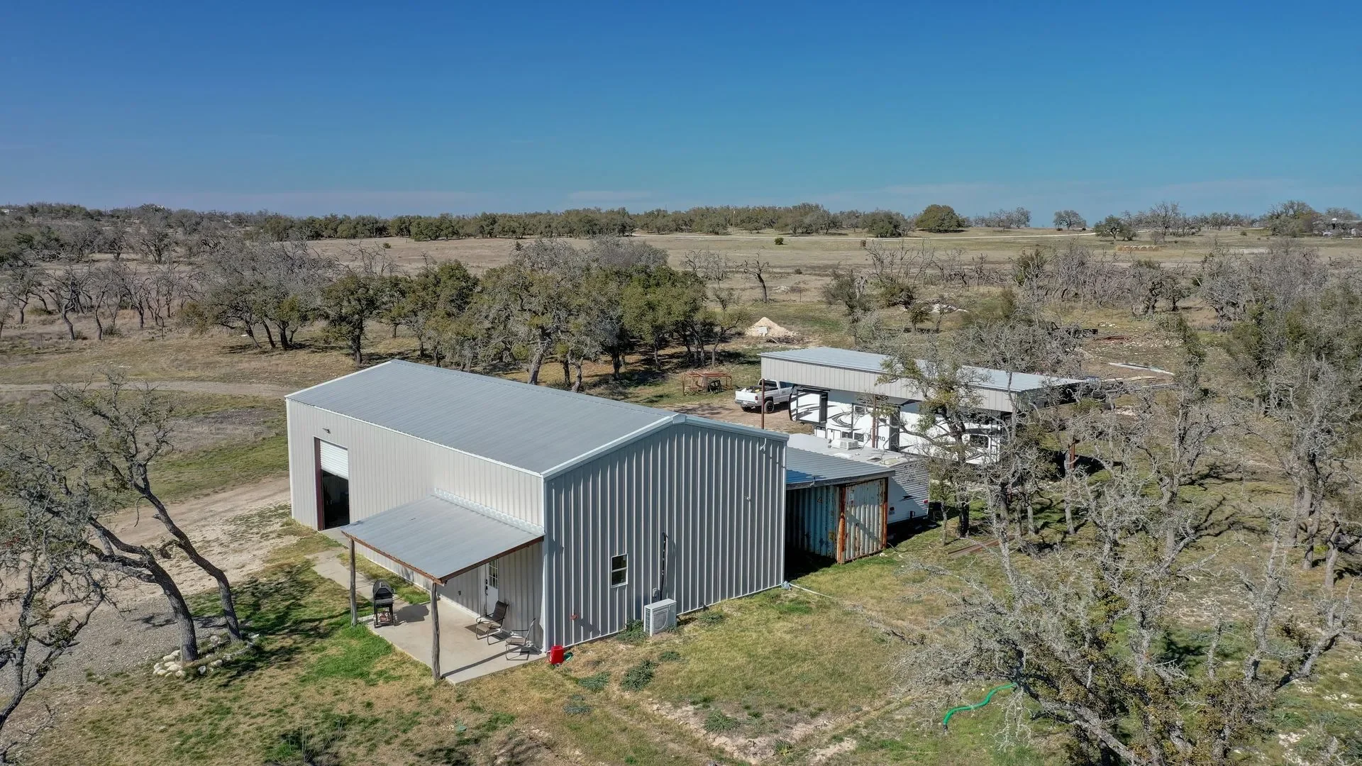 Aerial view of a farm with a large white metal barn, a smaller building with a porch, trees, and open fields under a clear blue sky.