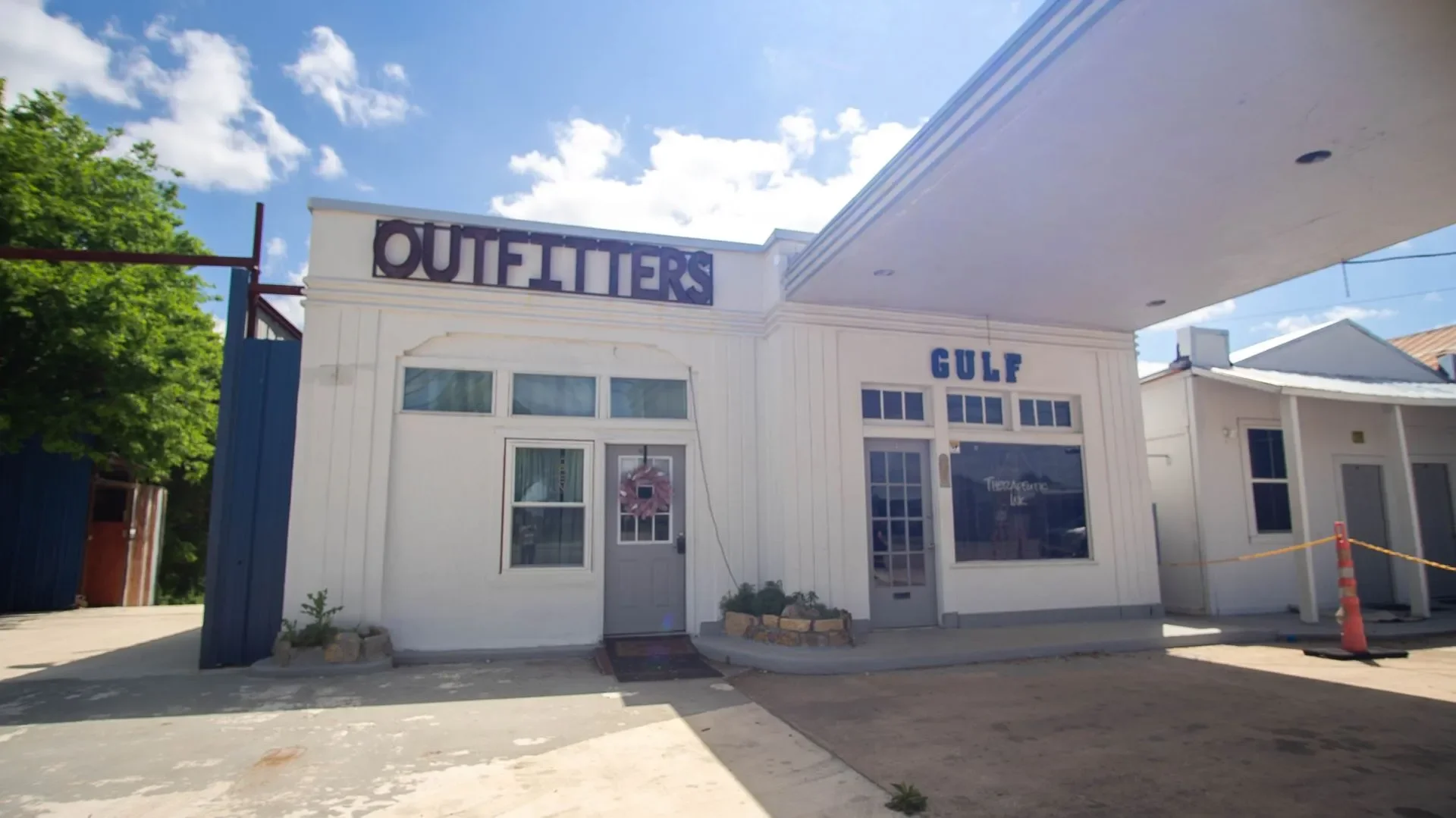 White building with signs reading 'OUTFITTERS' and 'GULF'; glass doors and windows, with a small potted plant outside, under a blue sky with clouds.