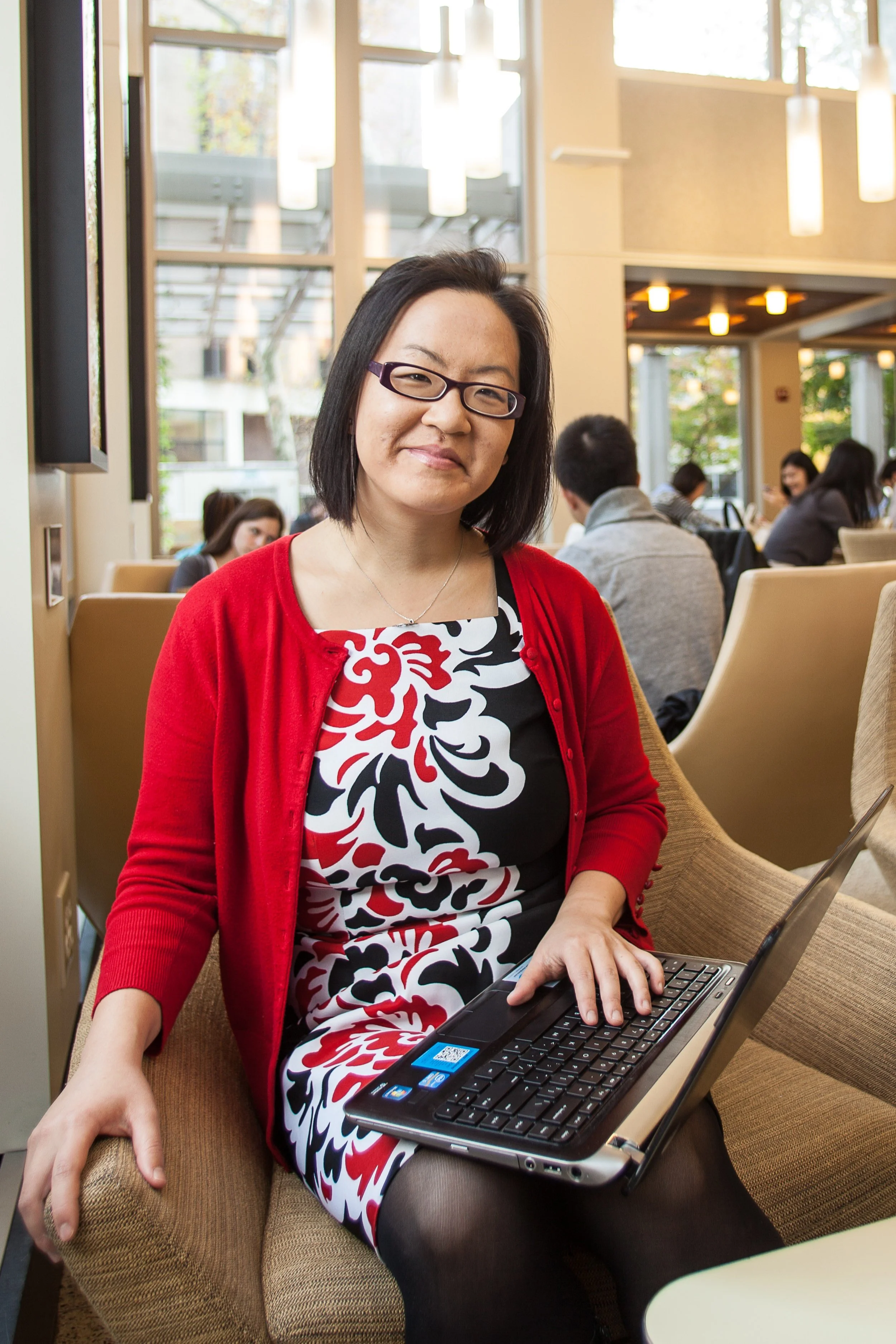 Woman with glasses sitting on a beige chair at a table, using a laptop in a bright, modern cafe with large windows and hanging lights.