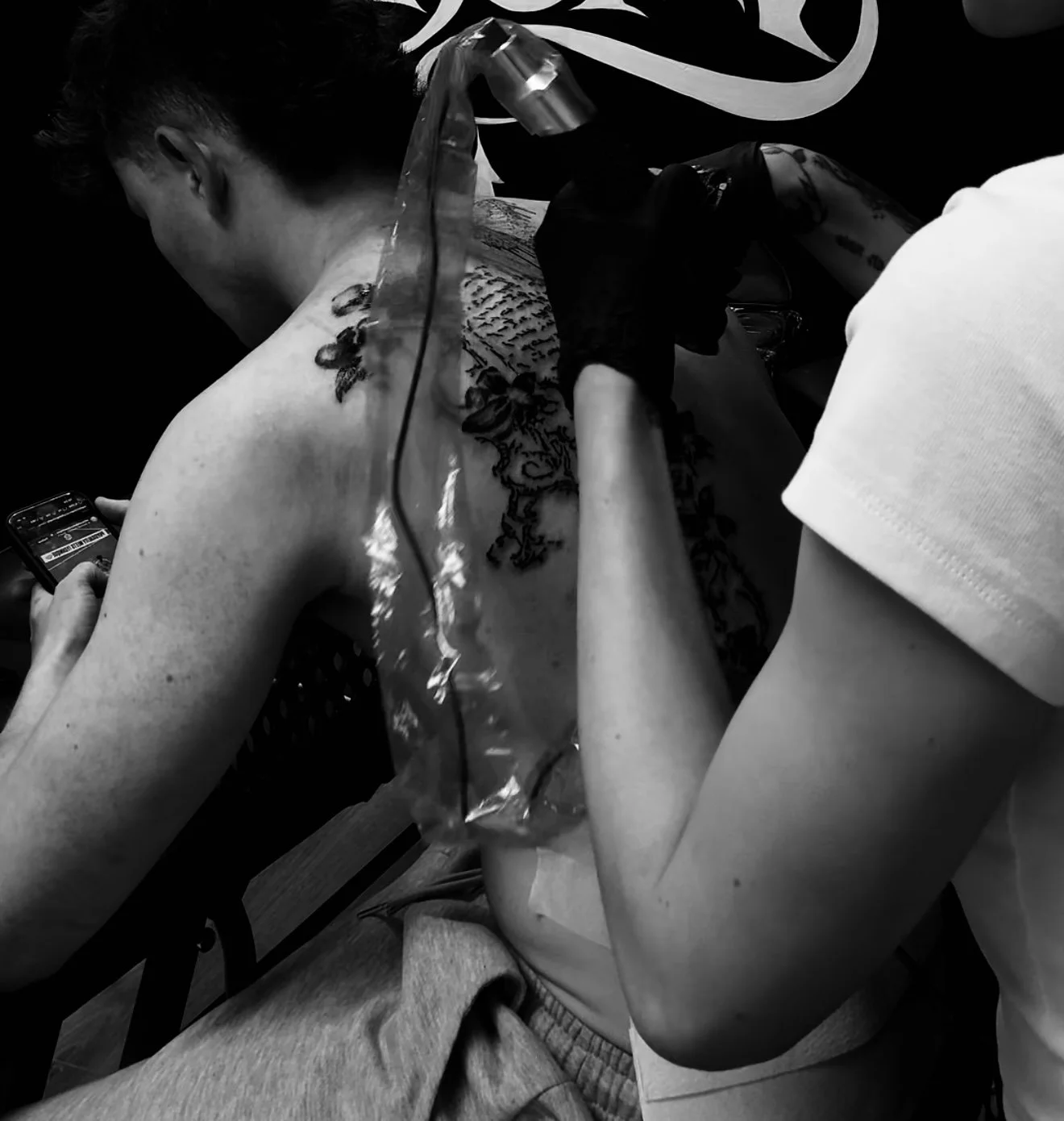 A person getting a tattoo on their upper back with a tattoo machine, while another person oversees the process. The person receiving the tattoo is looking at a mobile phone and has a floral tattoo on their shoulder. The scene is in black and white.