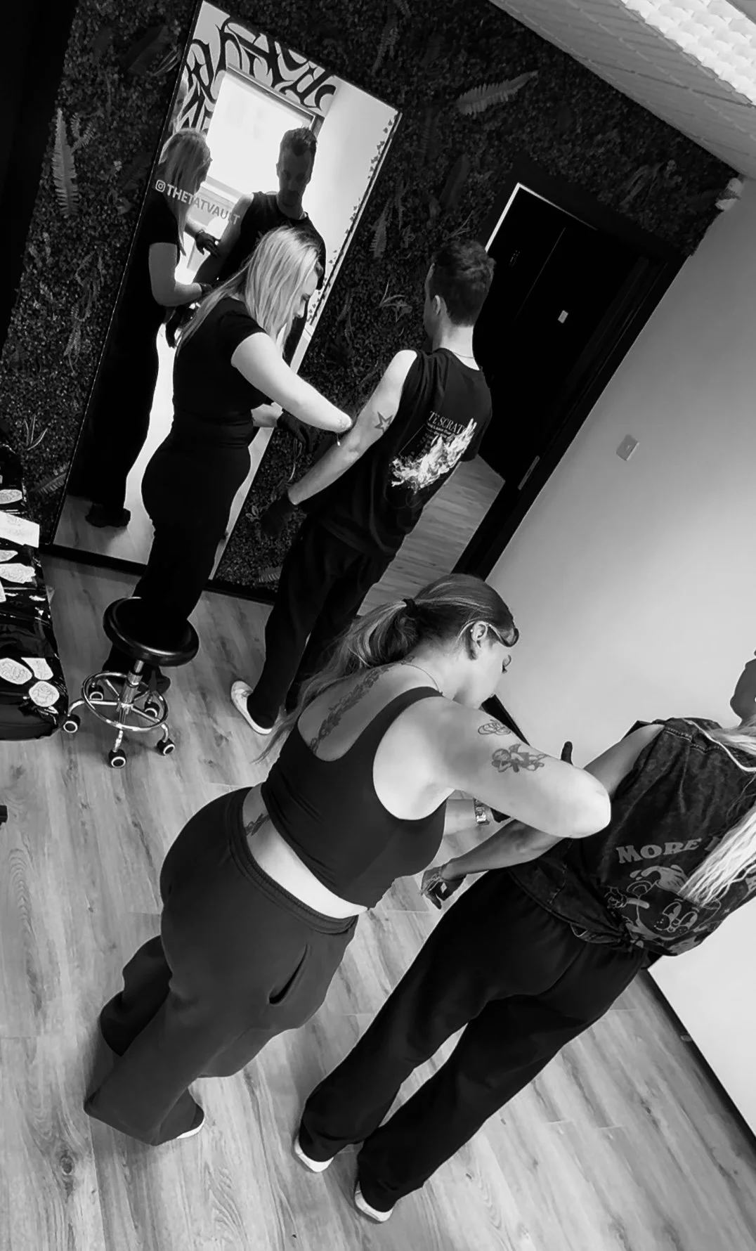 People getting tattoos in a tattoo studio, with one person seated and another actively tattooing. Two women are standing nearby, one with tattoos on her arms and back, and a third woman with blonde hair sitting in front of a mirror.