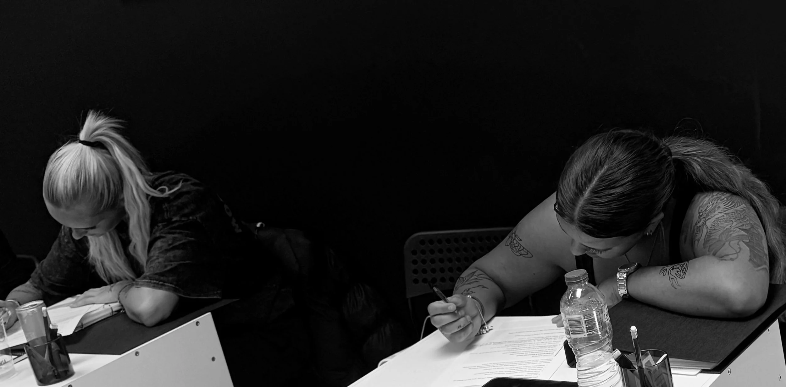 Two women with tattoos sitting at a table, writing on papers, with water bottles and pens on the table, against a dark background.