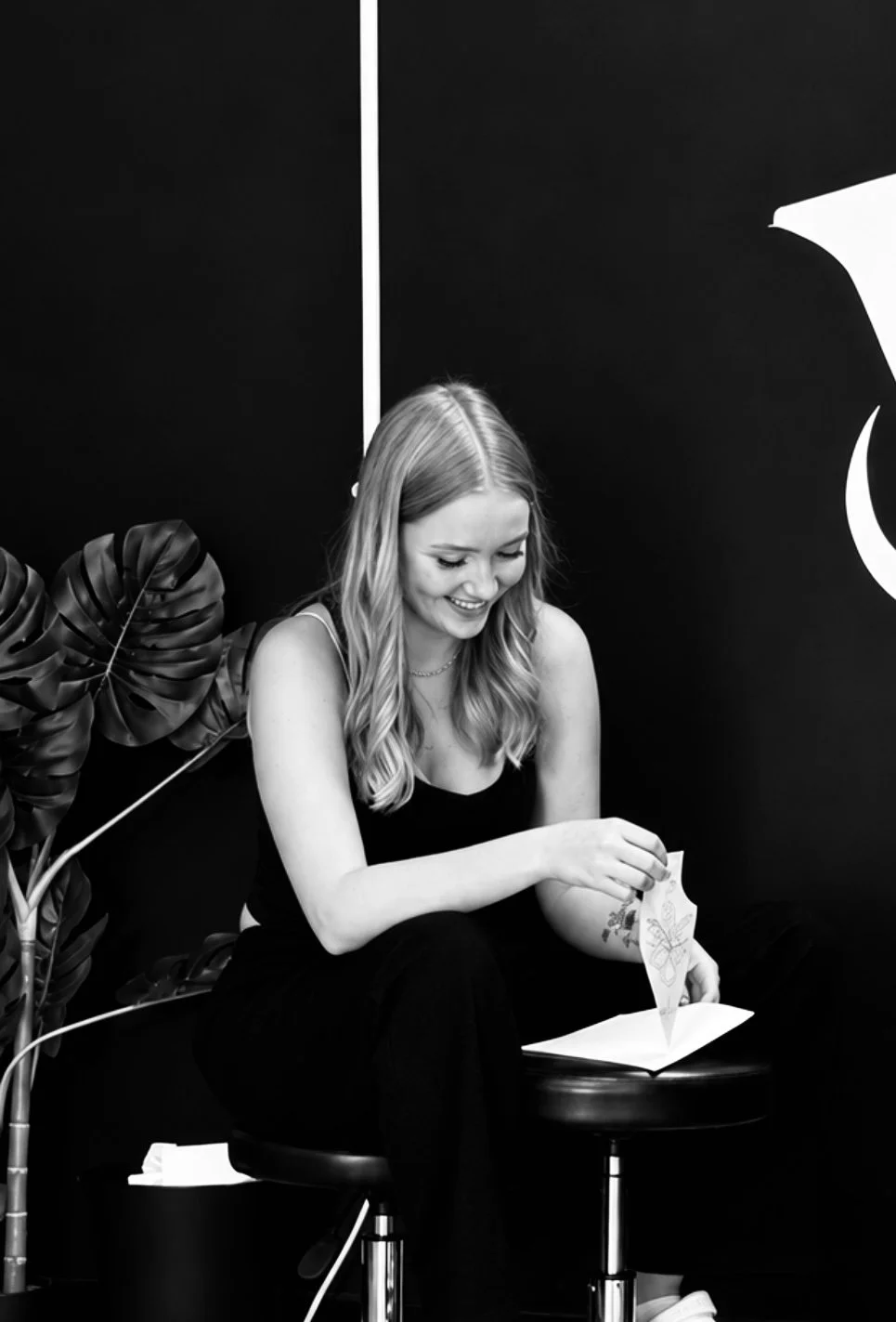 A young woman with wavy hair sitting on a stool, smiling and reading a letter or card in a black and white photo, with large leafy plant beside her and a dark background with a partial view of a sign or logo.