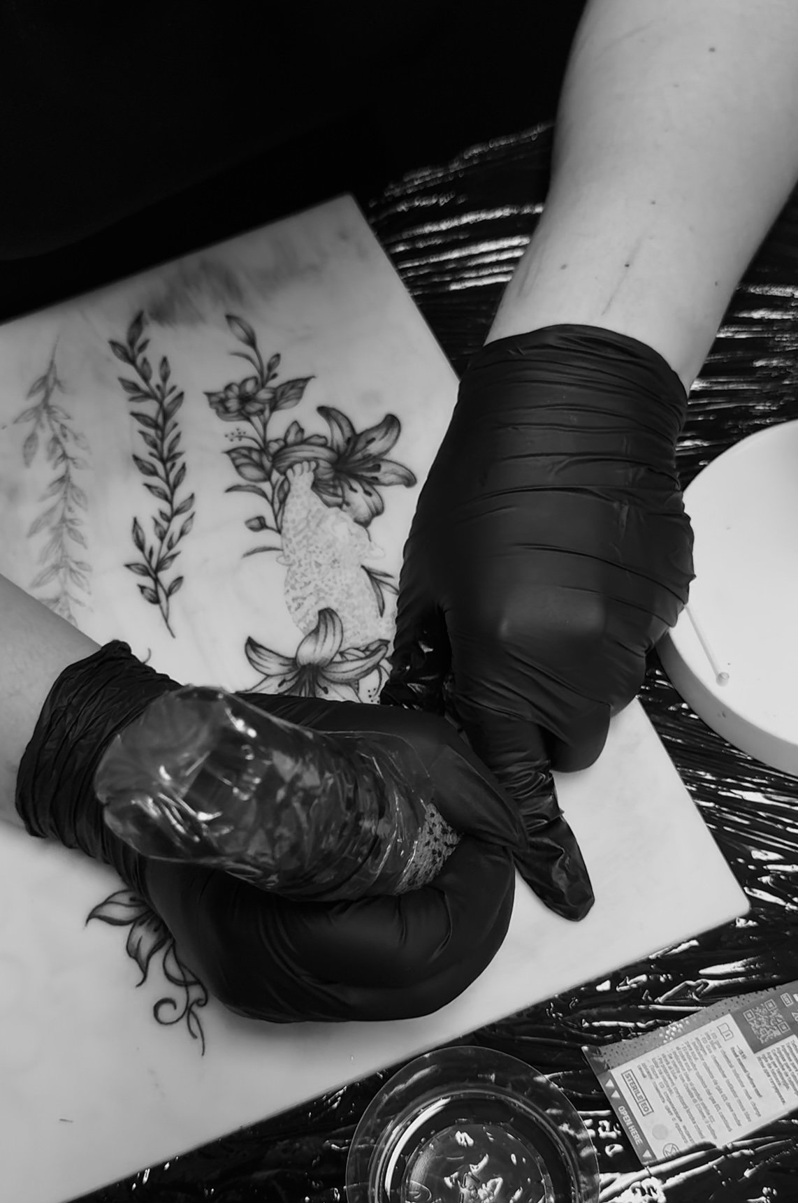 Tattoo artist in black gloves working on a floral tattoo design on a person's thigh using a tattoo machine.