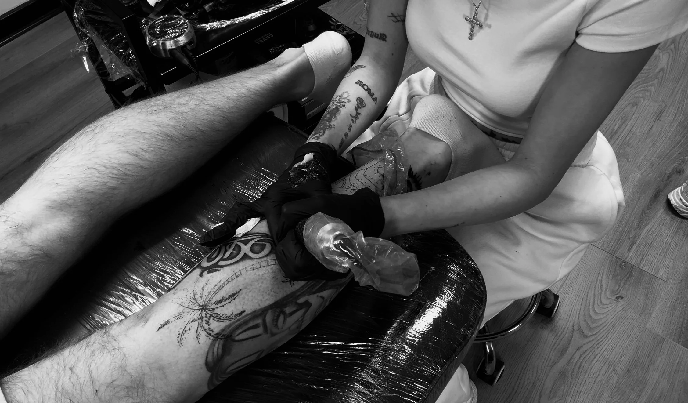 A person with tattoos on their arm sitting on a chair, receiving a tattoo on their forearm from a tattoo artist wearing gloves. The scene is in black and white.