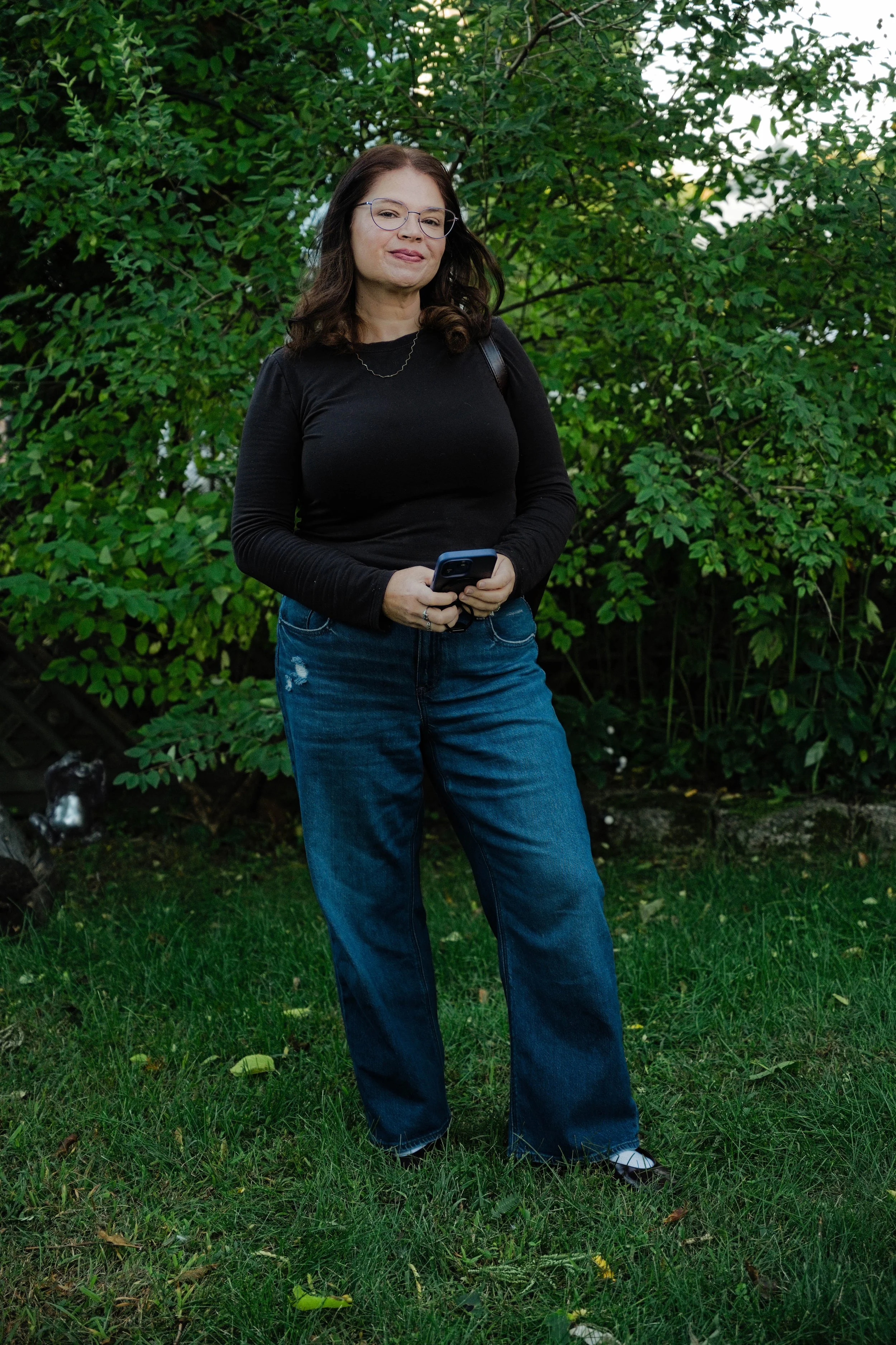 Sarah Aprea of Open Sign Socials standing outdoors in a Boston-area backyard, holding her phone