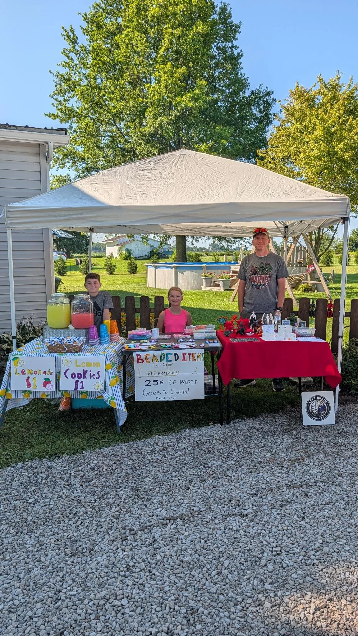 A family-friendly yard sale setup with a white canopy tent, three people standing behind the tables, and various items for sale, including lemonade, cookies, beaded items, and accessories. Signage indicates prices and that 25% of profits go to charity.
