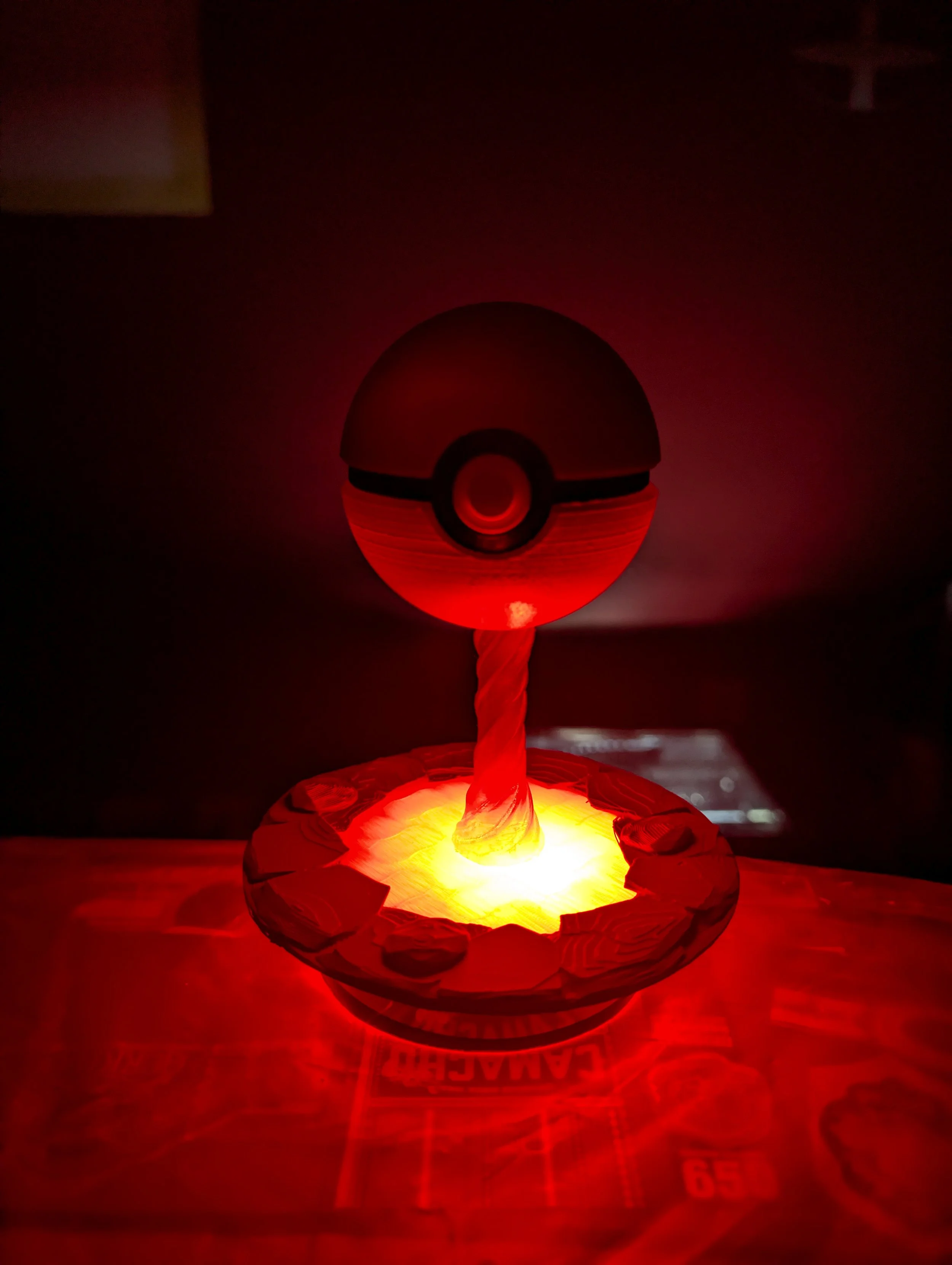A small glowing illuminated Pokéball figure on a cake with a candle on top, surrounded by a paper or plastic ring, casting a red light.
