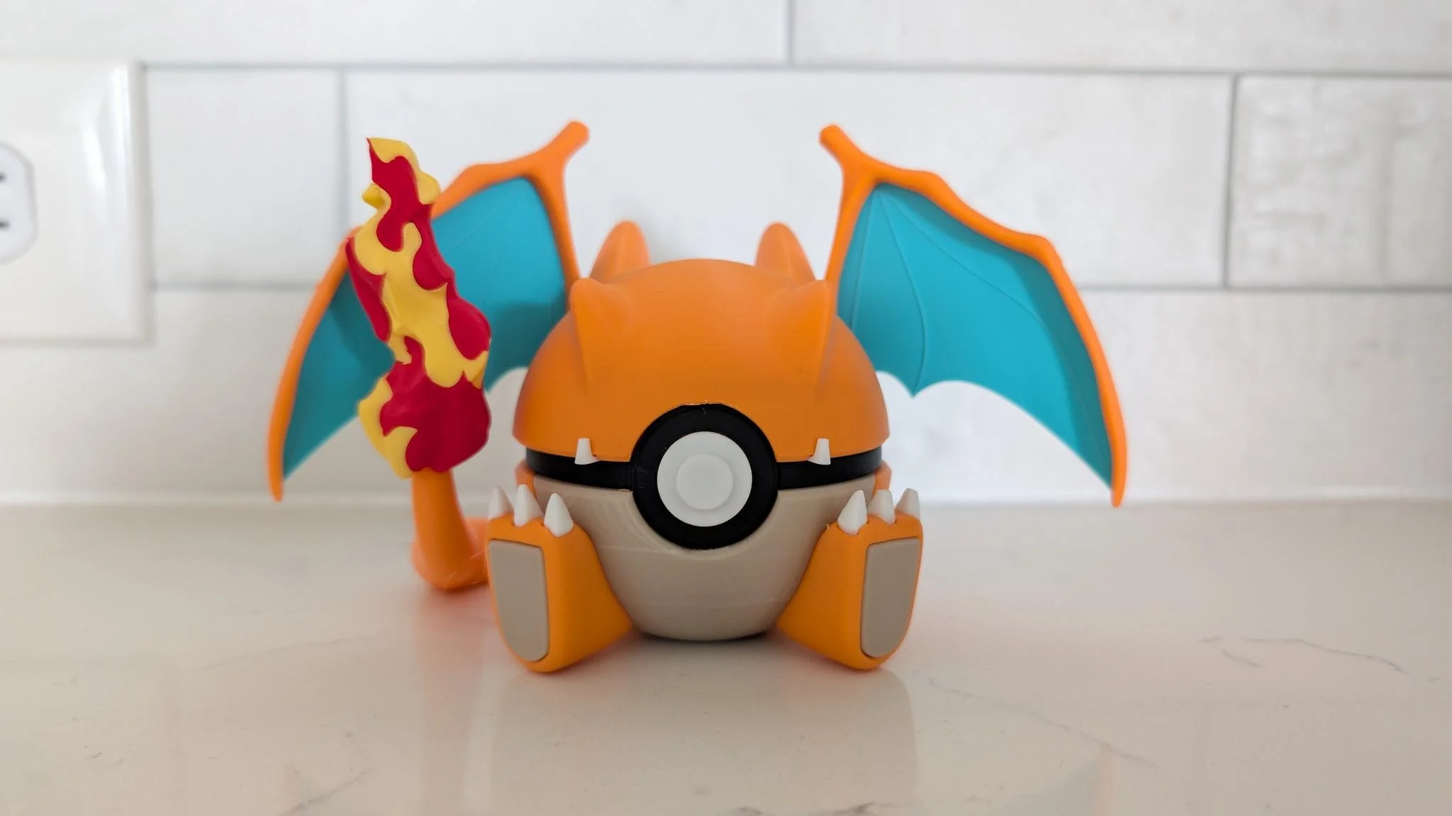 A toy Pokémon resembling Charizard, with orange body, blue wings, flames on the tail, and a Pokéball design on the front, placed on a white surface.
