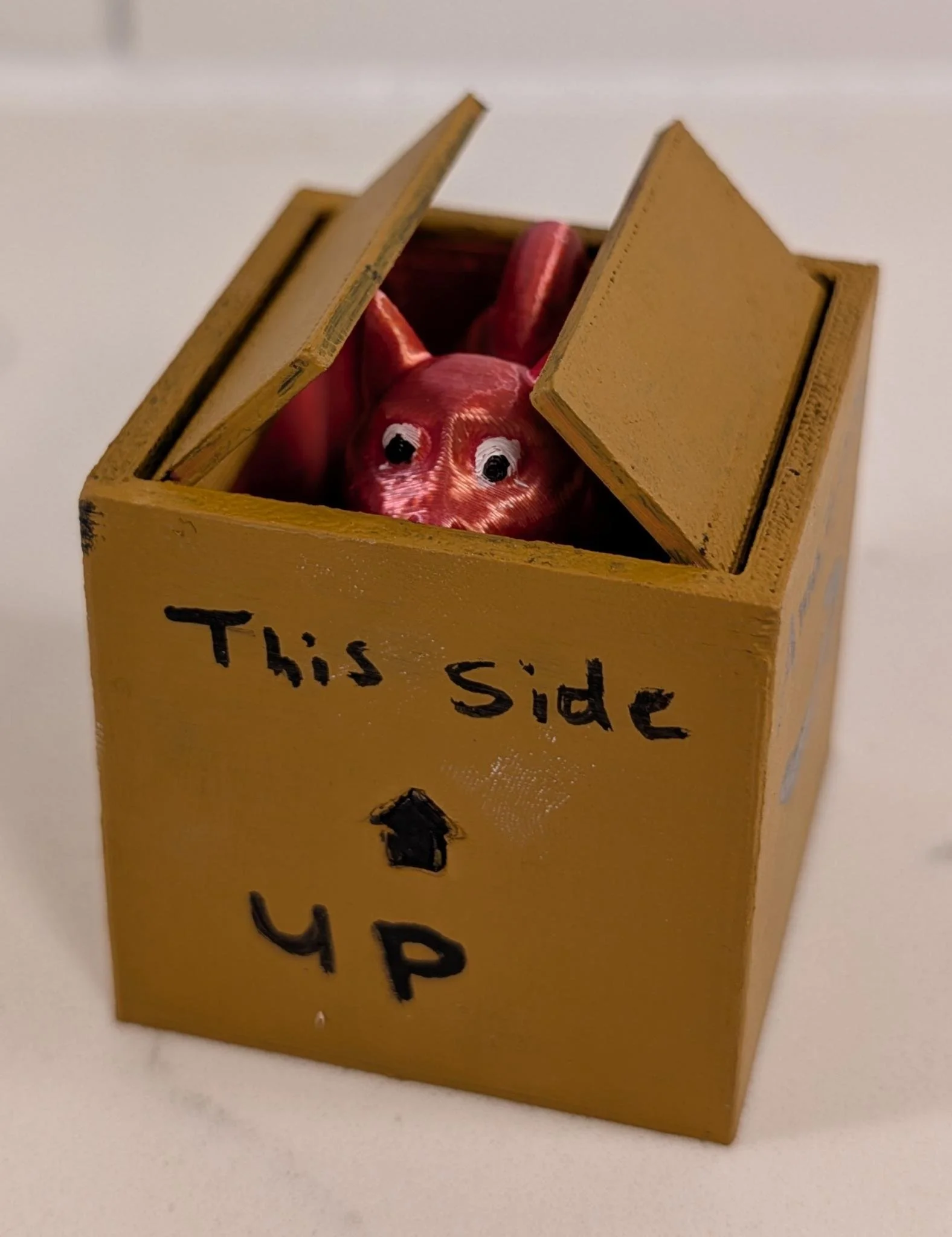 Small yellow cardboard box with the words 'This Side UP' written on it, containing a red, toy pig with big black and white eyes.