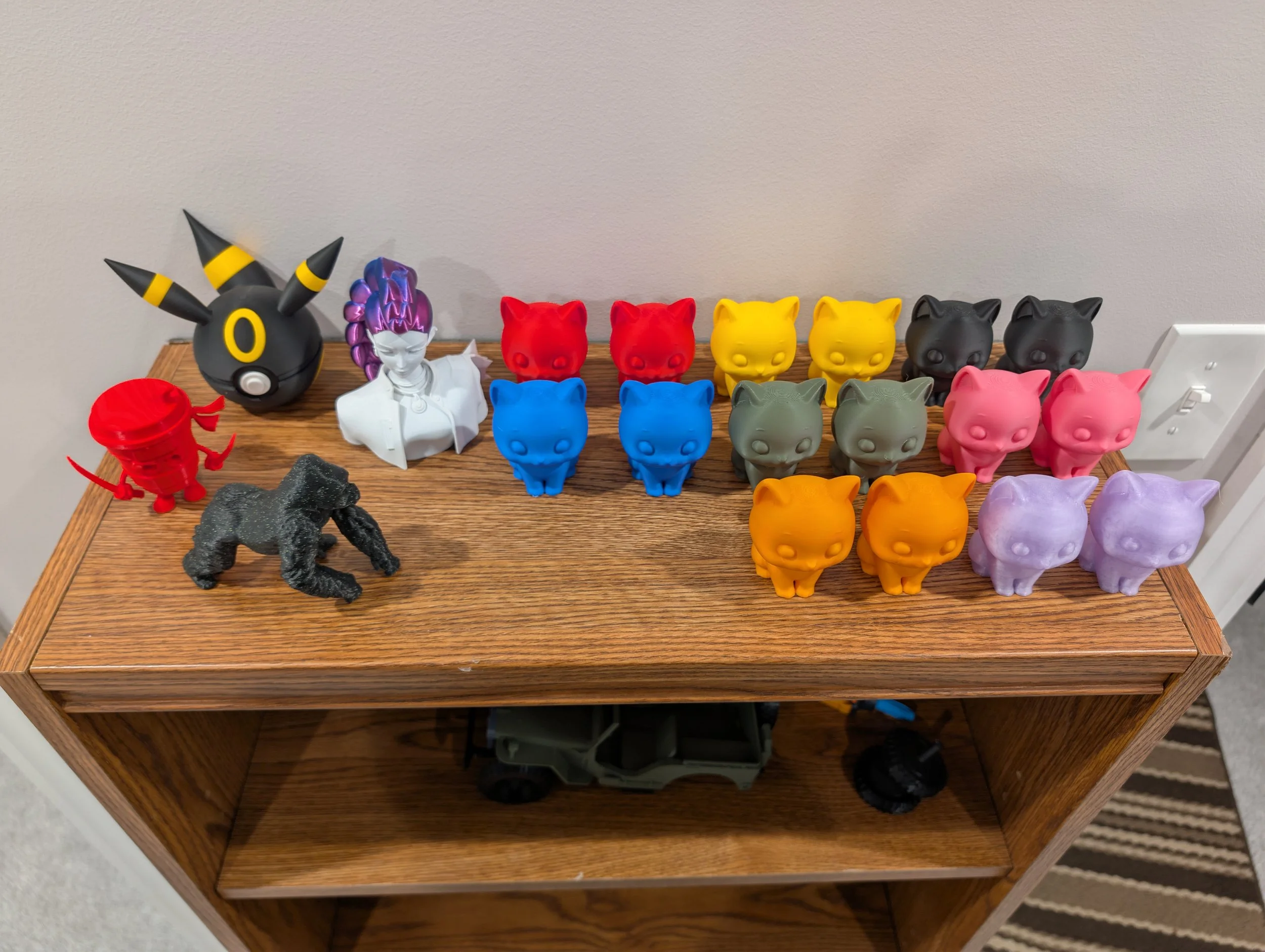 A wooden display shelf holding various colorful figurines and sculptures, including a large black and yellow creature on the left, a white female bust, several animal-shaped figurines in red, blue, yellow, gray, pink, and purple, and some other dark-colored sculptures on the left.