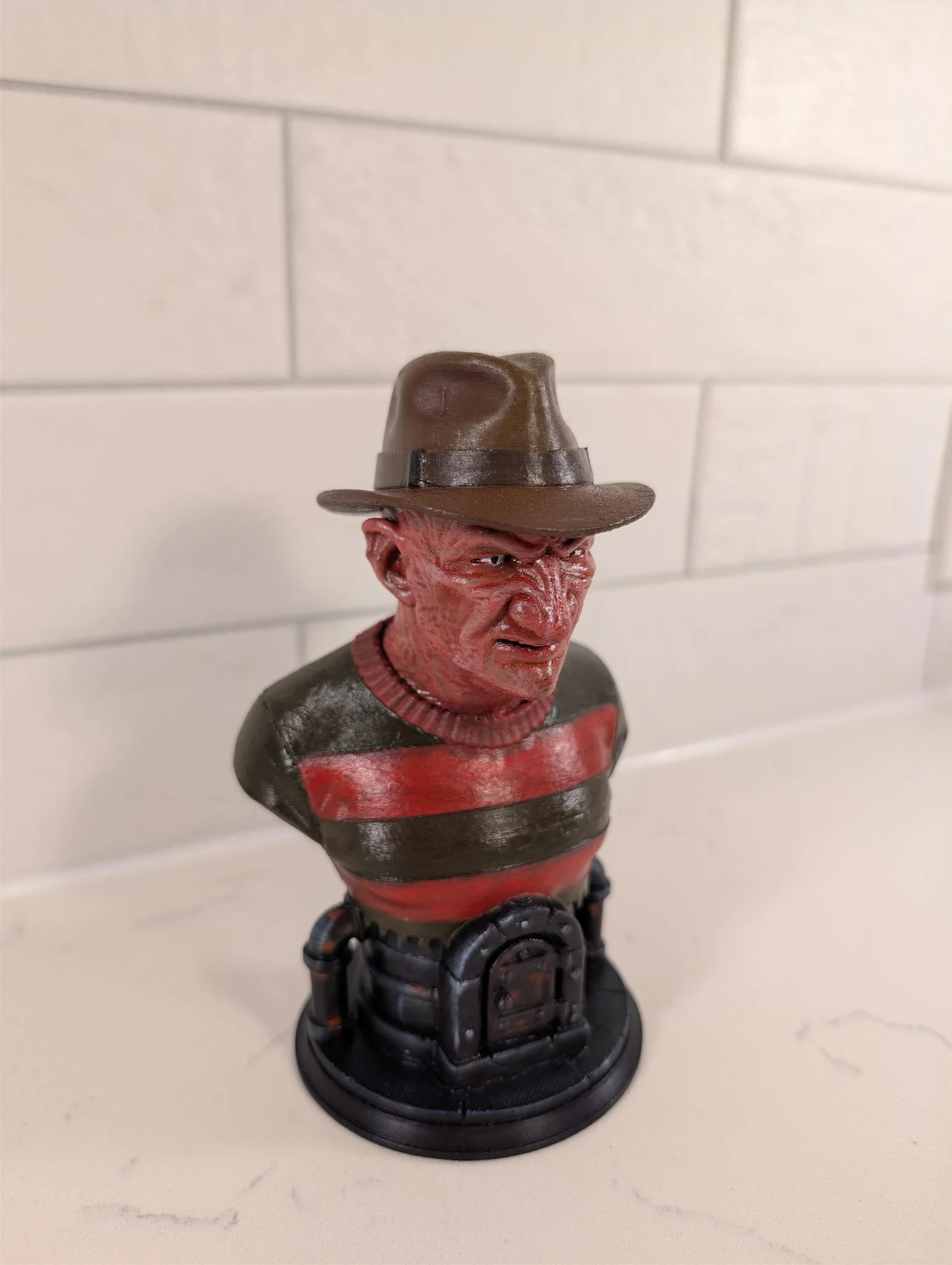 A figurine of Freddy Krueger from the horror movie series 'A Nightmare on Elm Street' wearing a brown fedora hat, with a red and green striped sweater, and a disfigured face, standing on a black circular base.