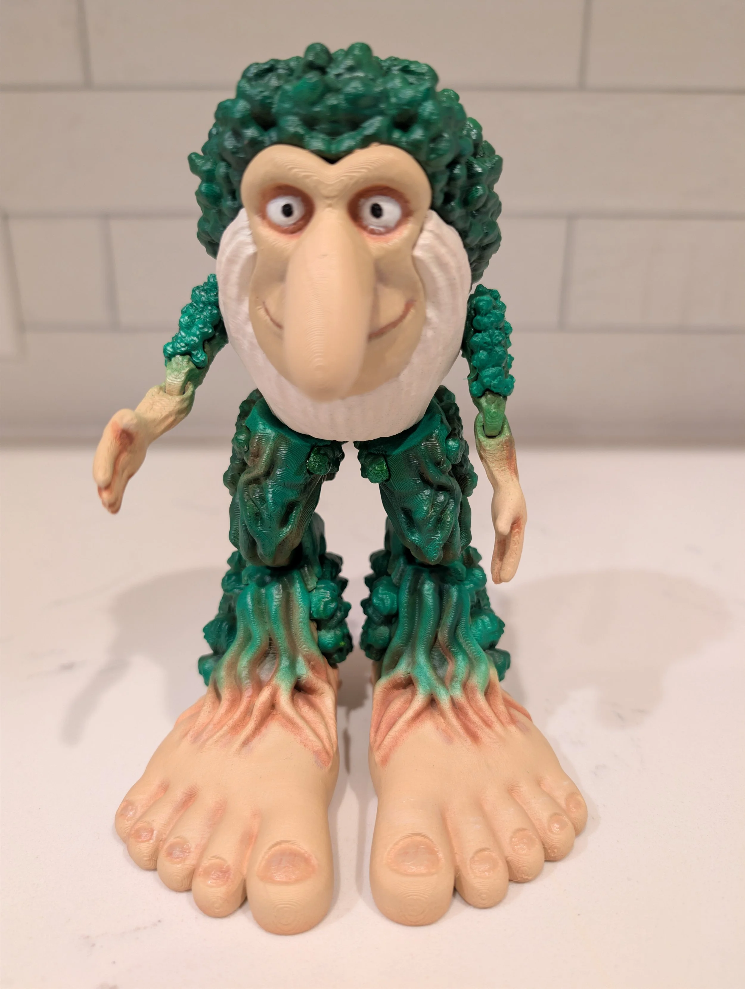 A toy figurine of a person with green curly hair, a long nose, and a smiling face, standing on feet that resemble human toes, with a background of white brick wall.