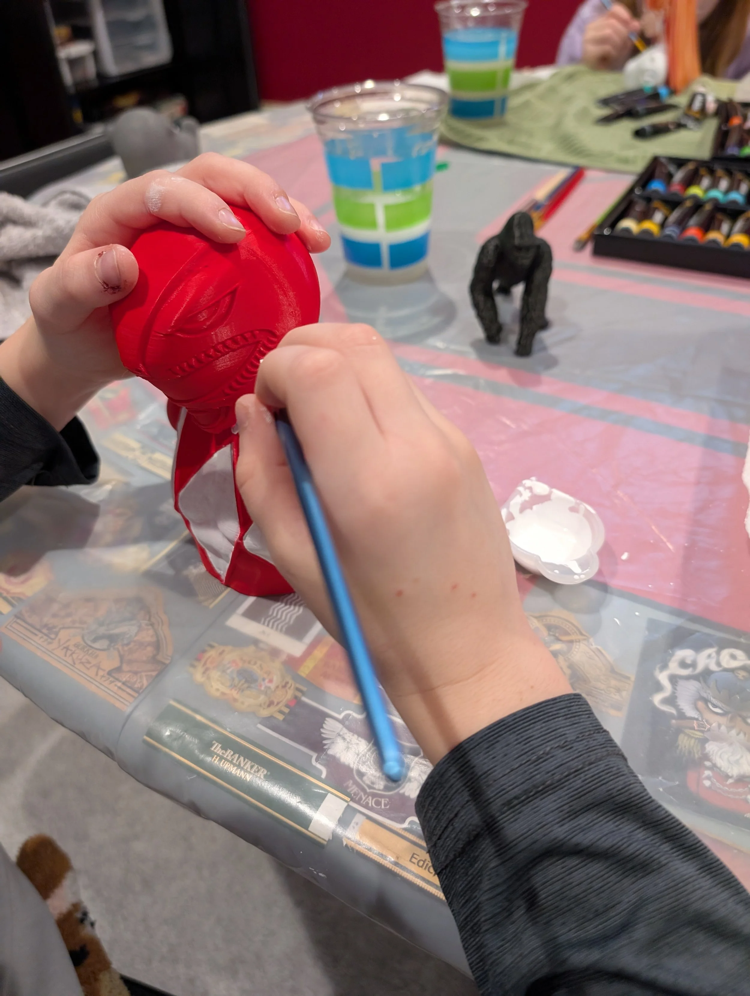 Child painting a small red figurine with a blue paintbrush on a table covered with a plastic tablecloth. There are two water cups, one with blue and green stripes, and a black toy gorilla. Art supplies and other children are in the background.