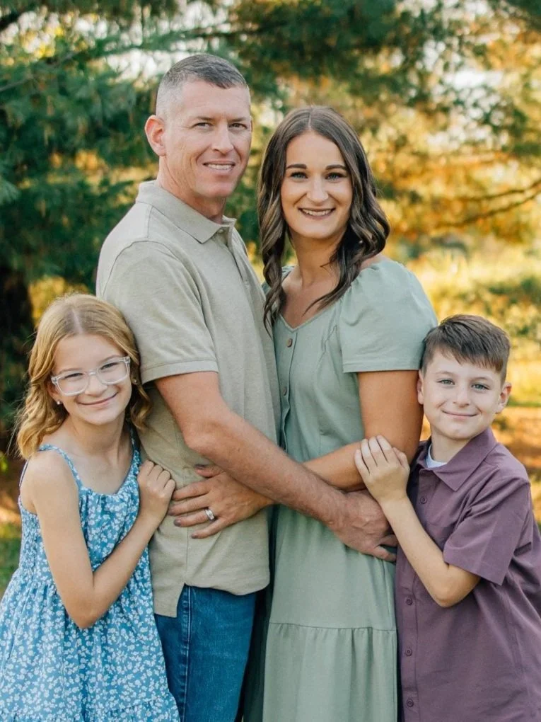 Family portrait of two adults and two children outdoors during daytime, surrounded by trees with autumn foliage.