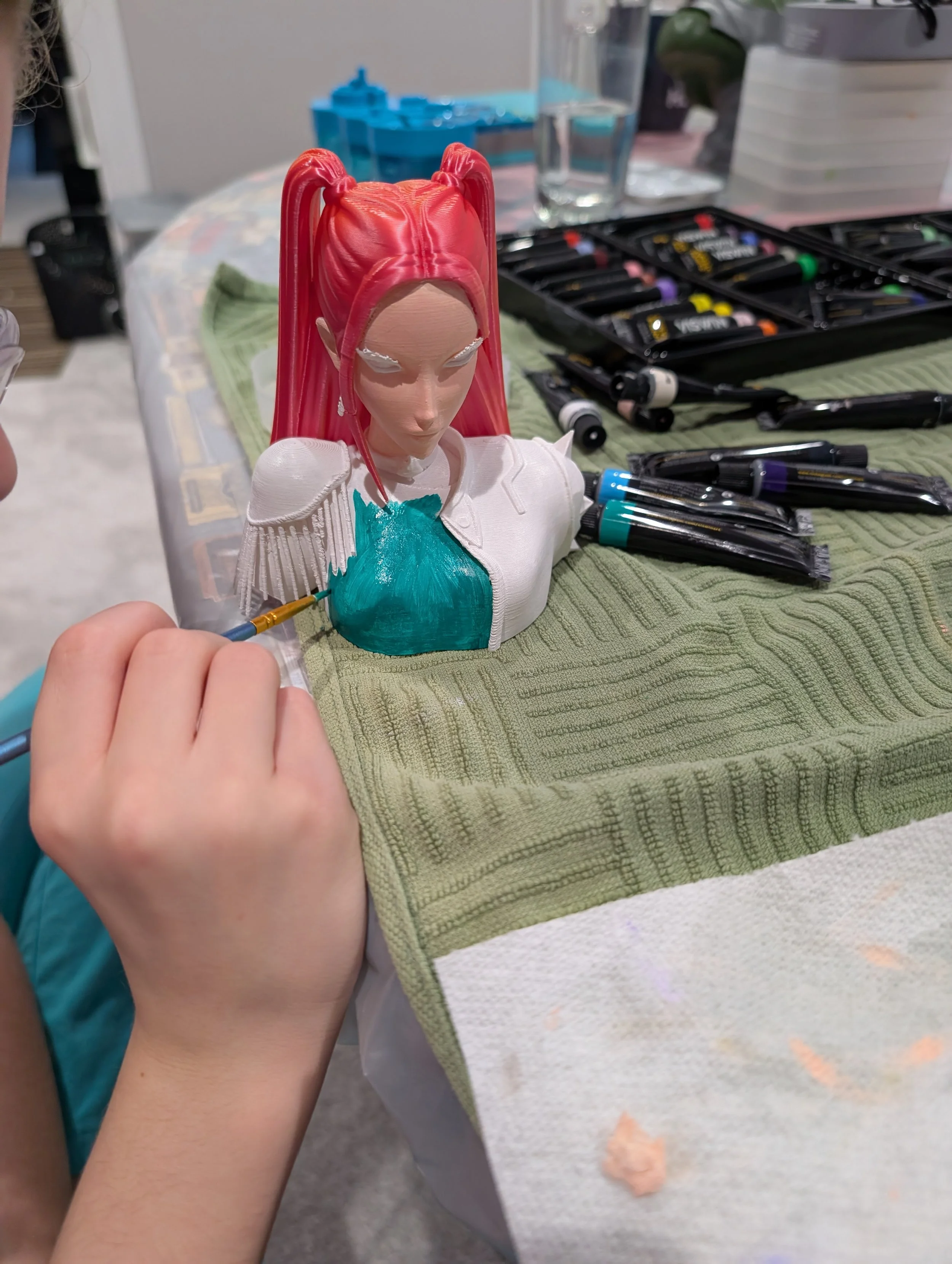 A person painting a figurine with pink hair and teal clothing. The figurine has an anime-inspired appearance and is placed on a green towel on a table with art supplies nearby.