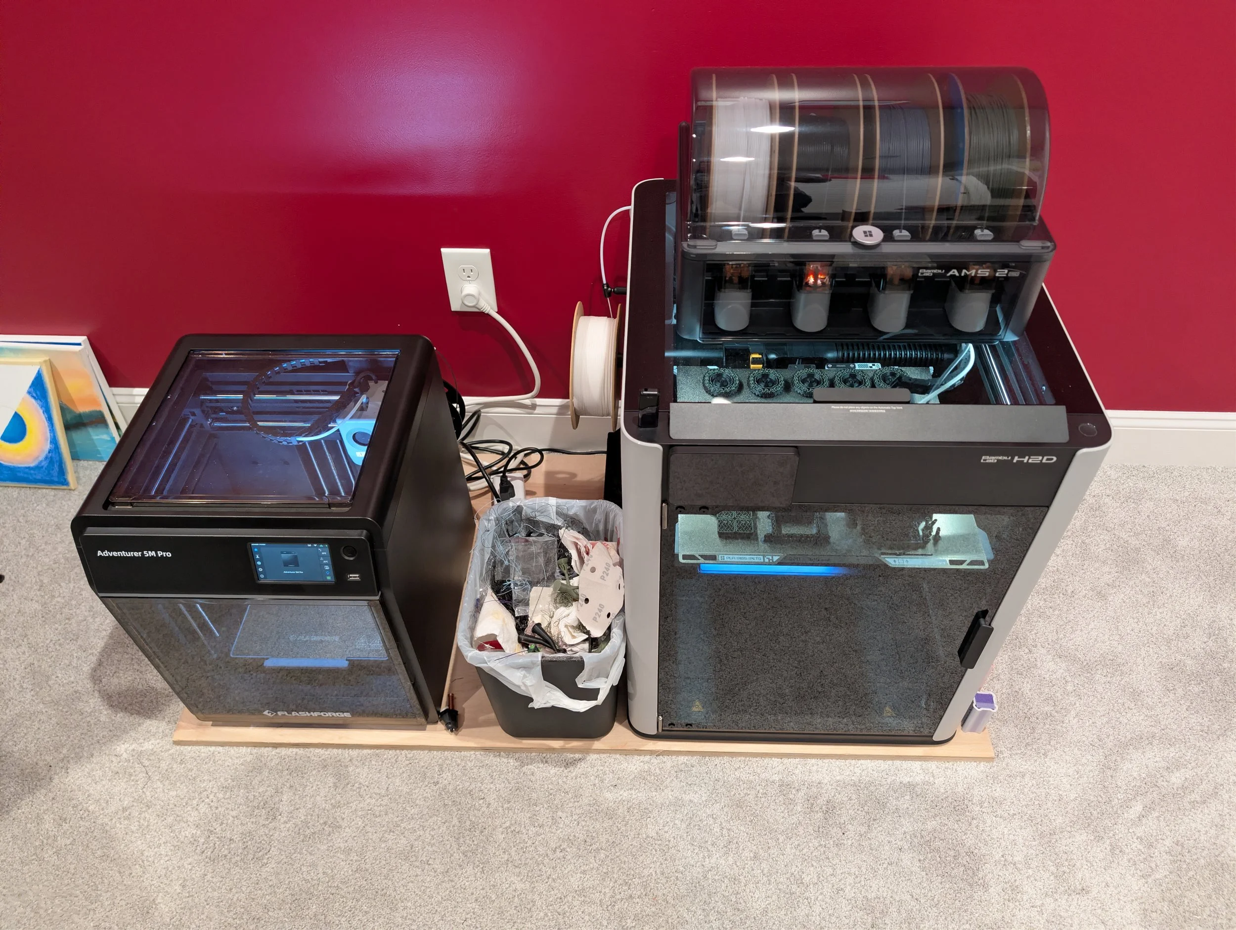 A red wall with two 3D printers on a wooden table, one in the foreground and the other in the background, and an abstract painting leaning against the wall.