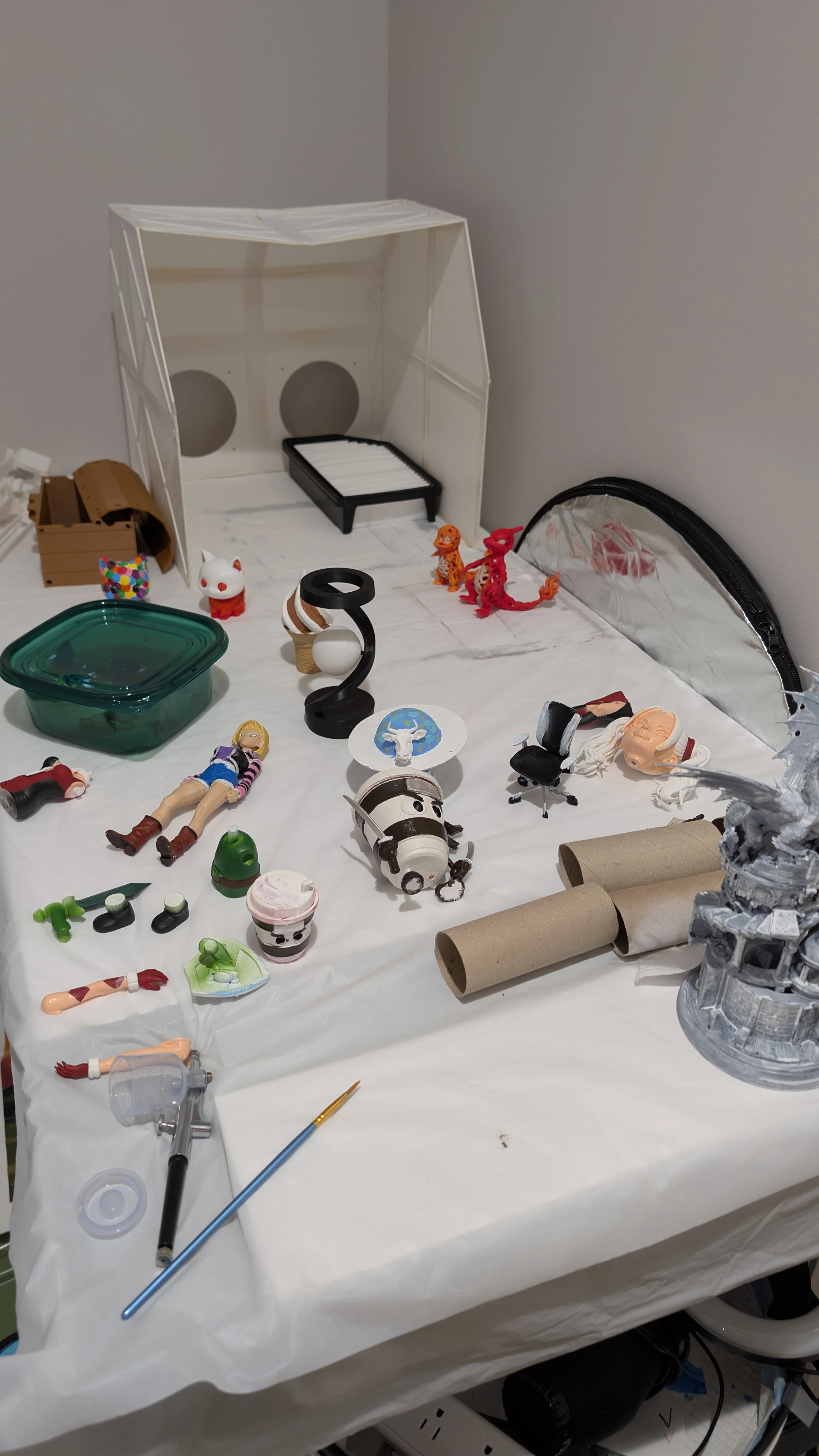 A cluttered work table covered with various toys, figurines, and crafting tools, including a small white animal figure, a toy horse, rolled paper, a small green container, and crafting paint. In the background, there is a white partition, some small plastic characters, and a reflective silver bag.