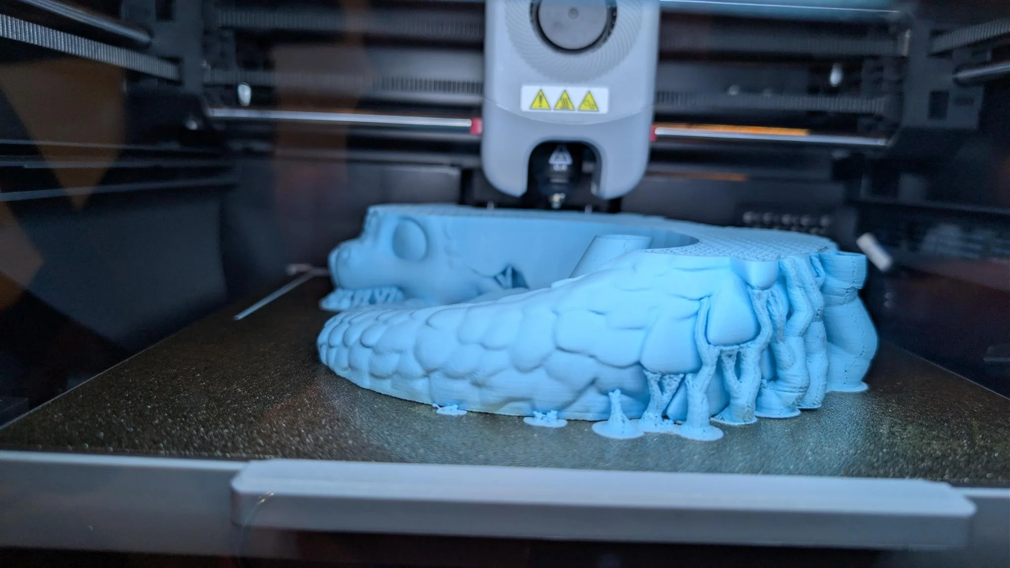 A 3D printer printing a blue model, which appears to be a creature with large jaw and teeth, inside the printer's enclosed chamber.