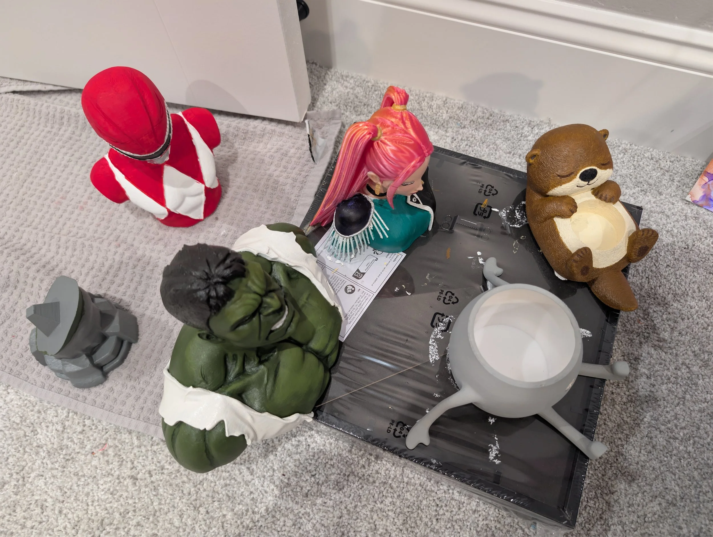 A collection of figurines including a red and white superhero, a pink-haired girl, a brown bear, a green Hulk figure, and a gray building, placed on a beige carpet near a white wall with a small hole.