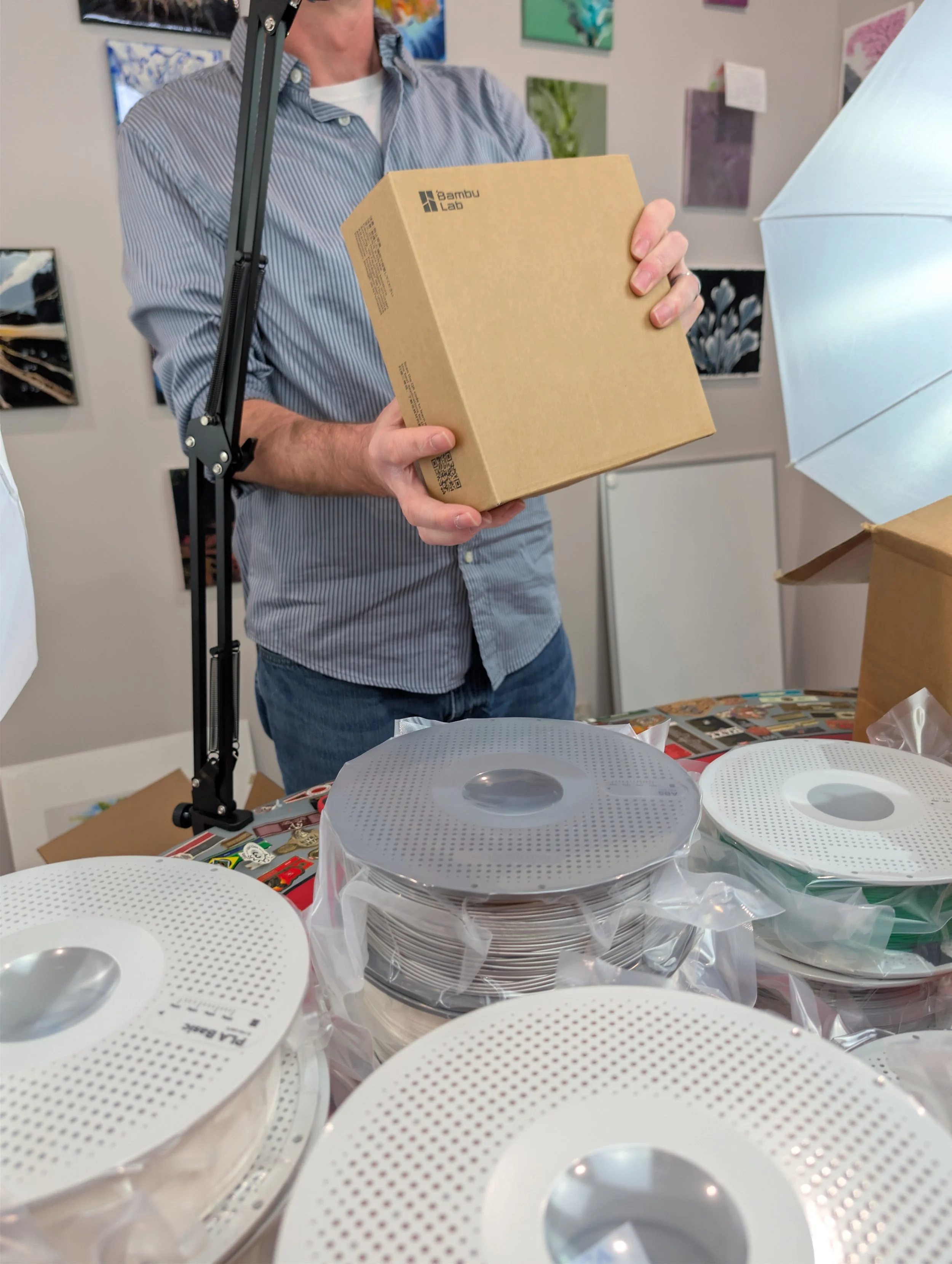 A person in a blue striped shirt holding a cardboard box with the label 'Bambu Lab' in a room with colorful wall art. Several rolls of white filament are on a table in the foreground, and a studio light is set up nearby.