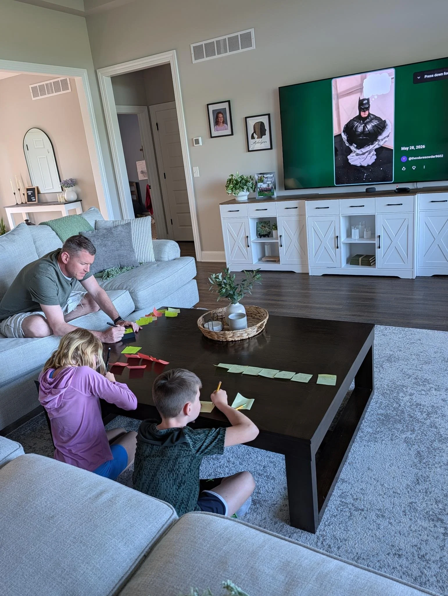 A living room with a large flat-screen TV on the wall showing a photo of Batman with a speech bubble, and a group of children and a man sitting around a dark wood table, working with sticky notes and pens.