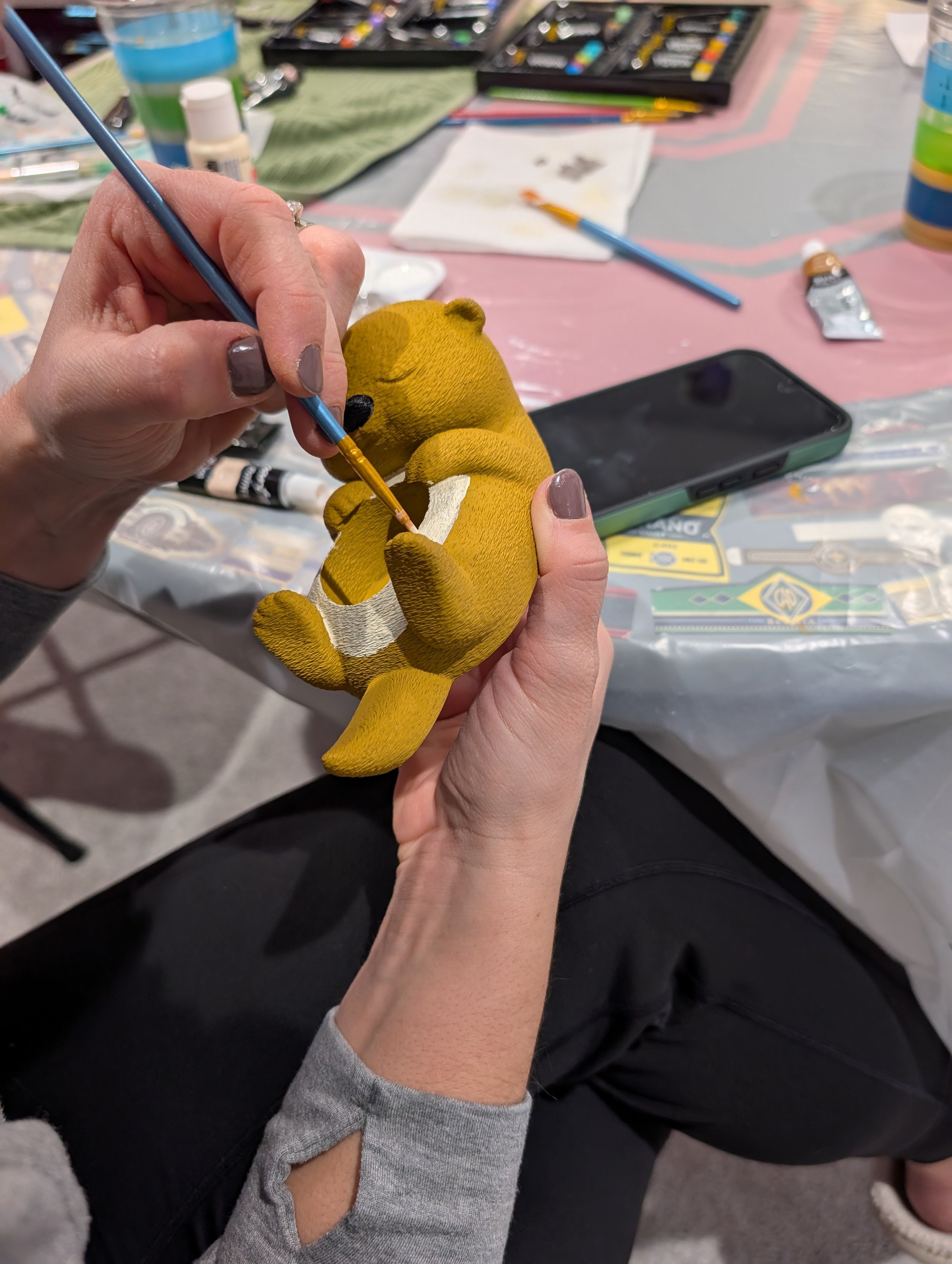 Person painting a small yellow plush toy with a paintbrush at a crafting table.