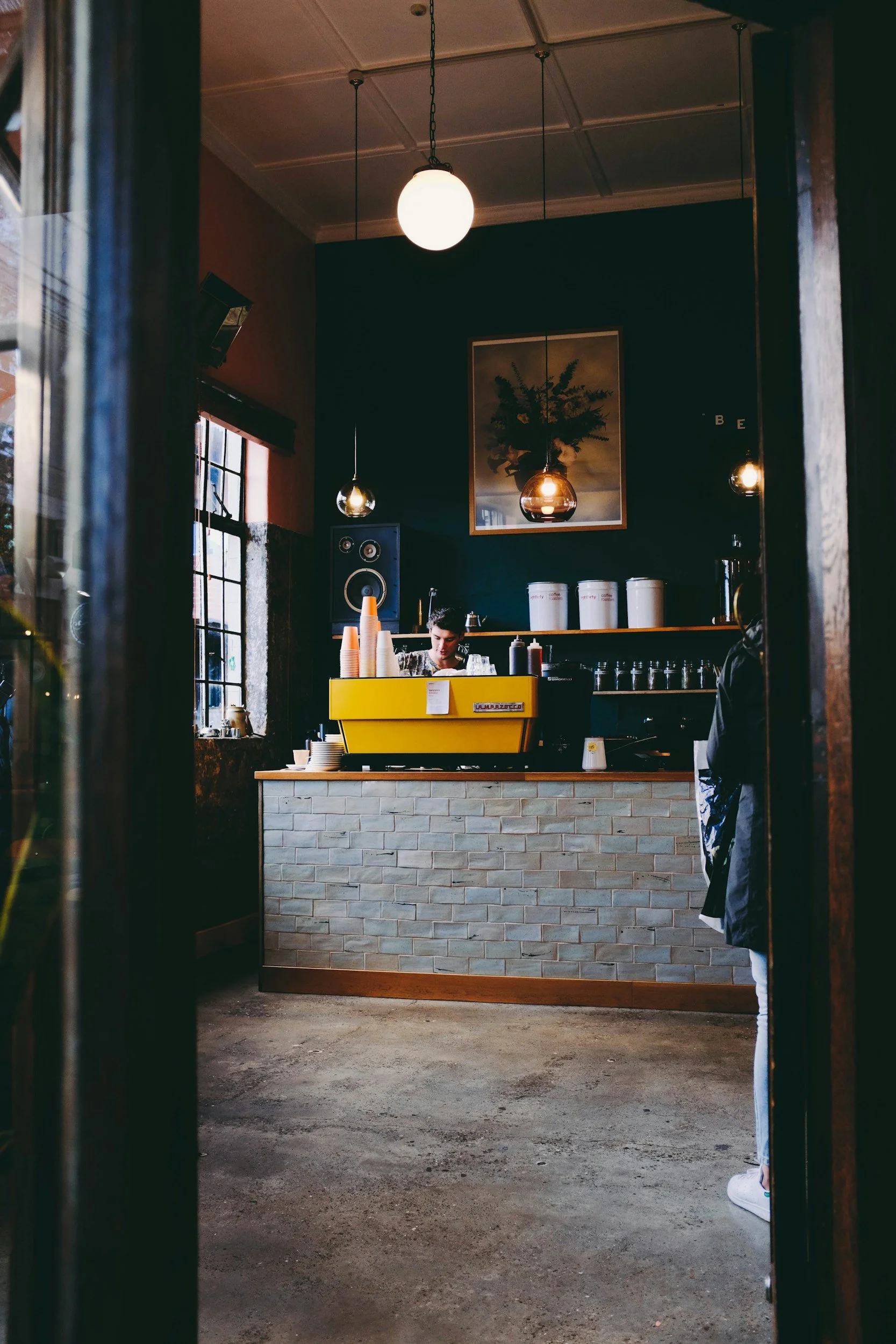 Inside a cozy coffee shop with dark green walls, hanging pendant lights, and a framed botanical picture. A barista is working behind the counter with cups, jars, and a yellow espresso machine. A person is partially visible on the right, wearing a dark jacket and patterned pants, standing near the counter.