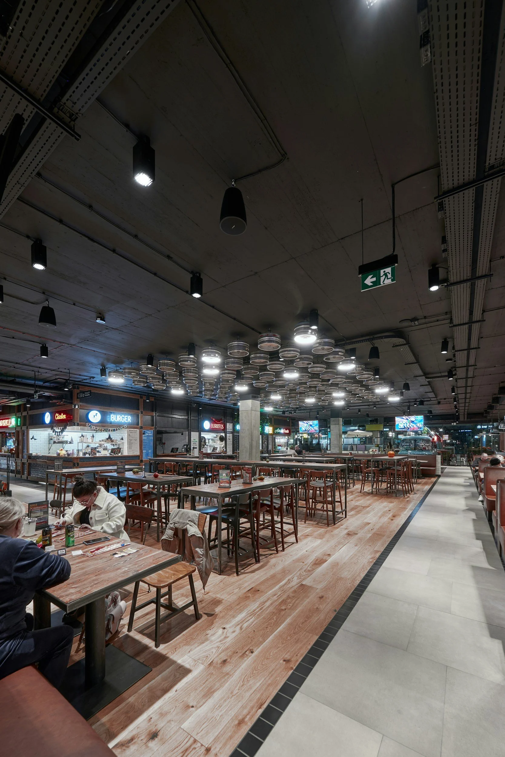 Interior of a modern food court with wooden flooring, multiple tables and chairs, restaurant counters displaying food brands like Burger and Burritos, and a ceiling decorated with circular light fixtures. An emergency exit sign is visible hanging from the ceiling.