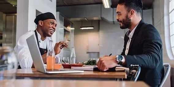 Two men, one in a chef's outfit and the other in business attire, sitting at a table and enjoying a conversation in a modern office or cafe setting.