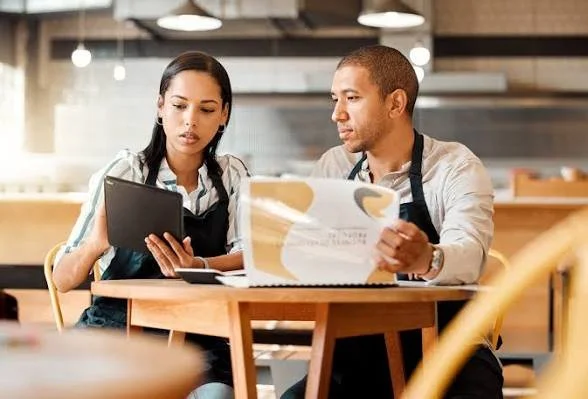 Two restaurant employees, a woman and a man, reviewing a menu or tablet at a table inside a restaurant.