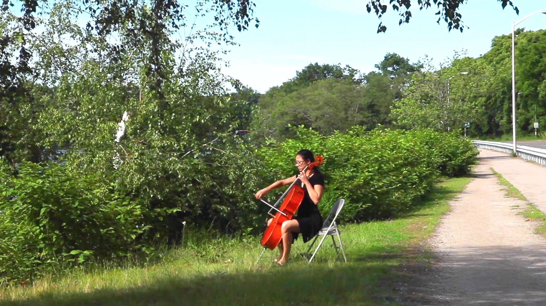 Cellist playing a cello outdoors beside a tree-lined path and road
