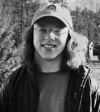 Black-and-white portrait of a smiling person wearing glasses, a baseball cap, and a jacket outdoors