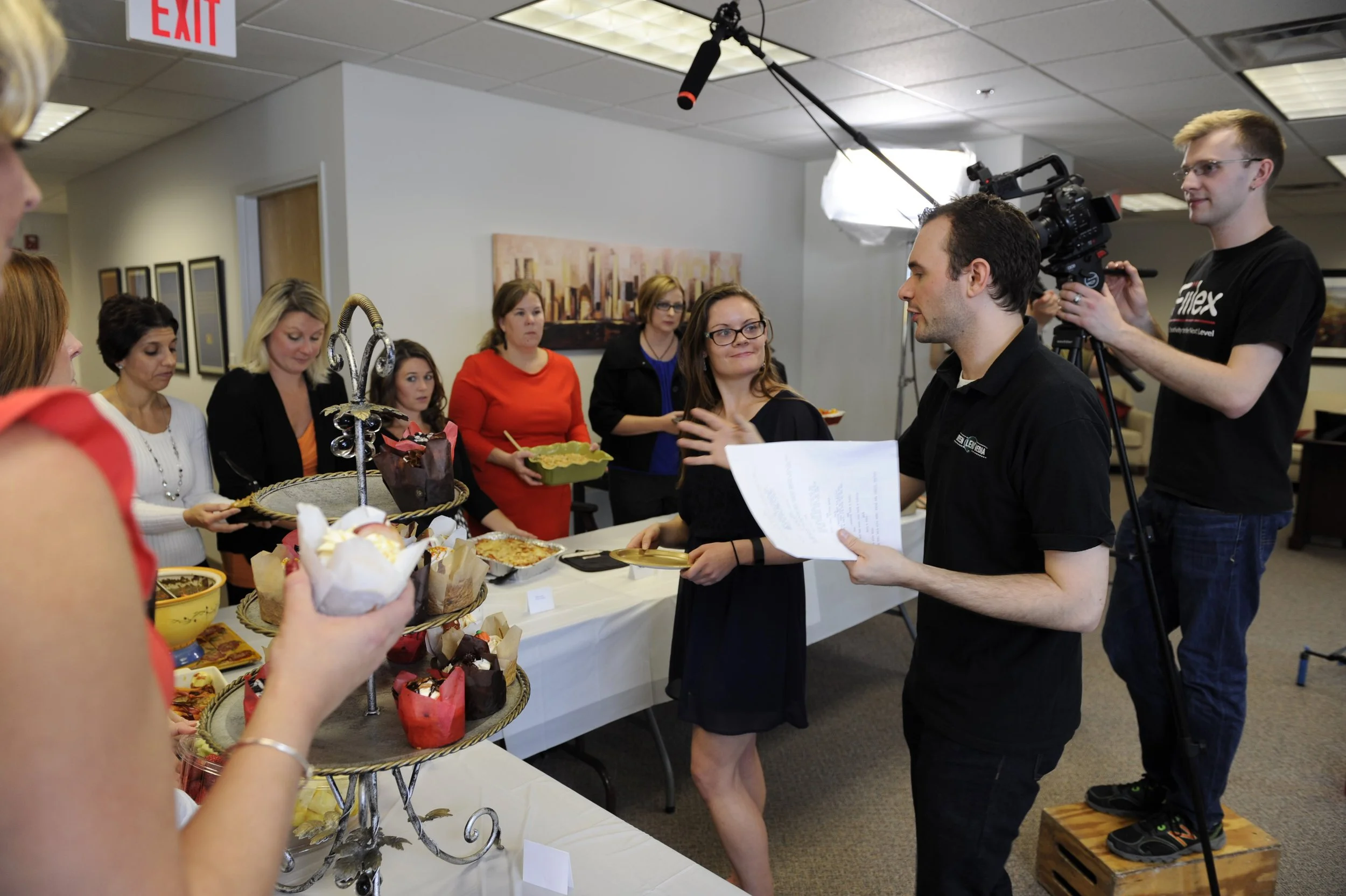 People gathered around a dessert table while a videographer films an office event.