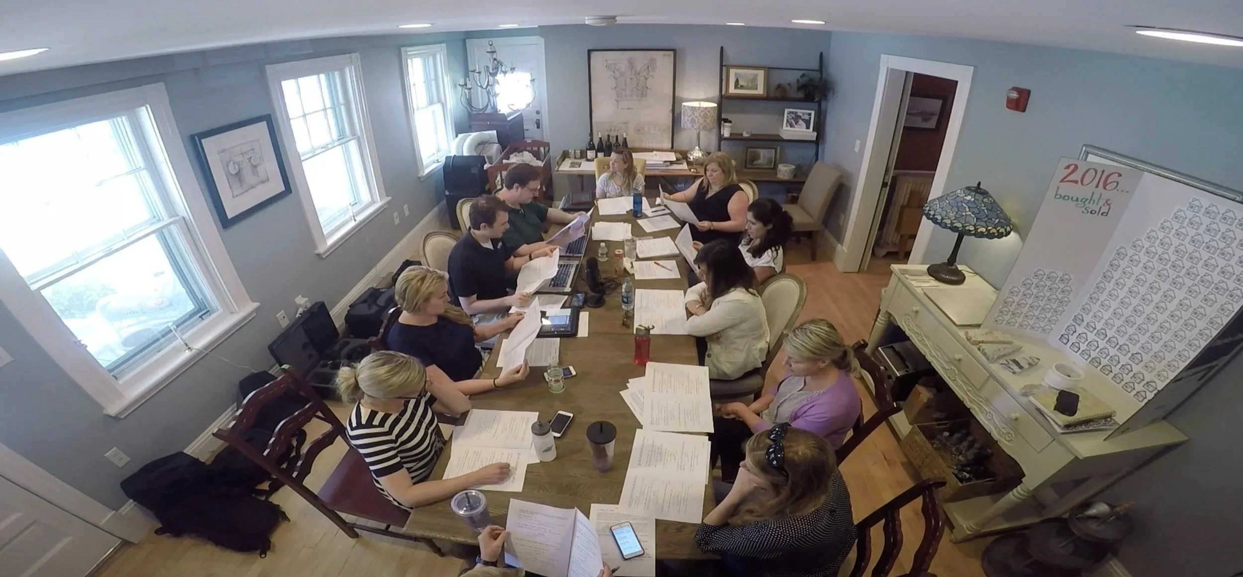 Group of people reviewing documents around a conference table in a bright office, with a “2016 bought & sold” board and lamp nearby.